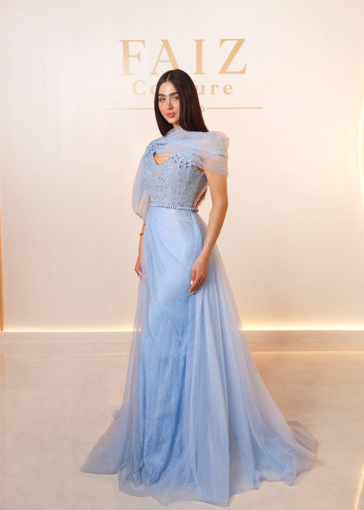 A woman modeling a light blue evening gown with sheer draped sleeves and a heavily embellished bodice, FAIZ Couture.