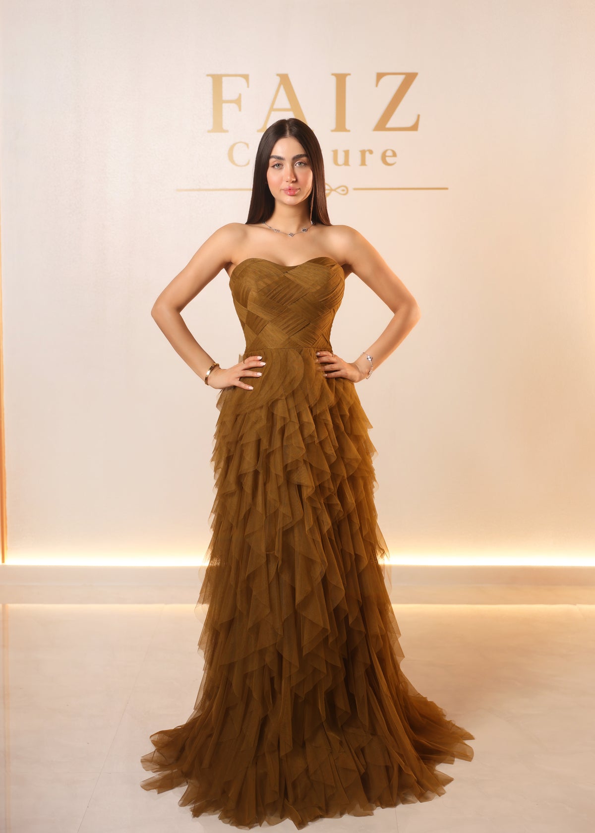 A woman stands with hands on hips wearing a strapless brown tulle gown with a structured bodice and layered ruffle skirt, FAIZ Couture