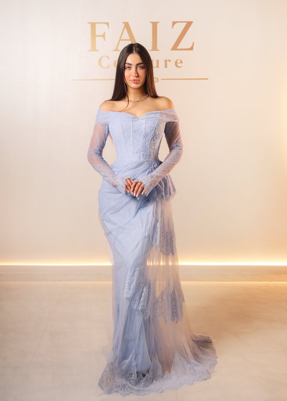 A woman models an off-the-shoulder light blue lace and tulle evening gown with long sheer sleeves, FAIZ Couture