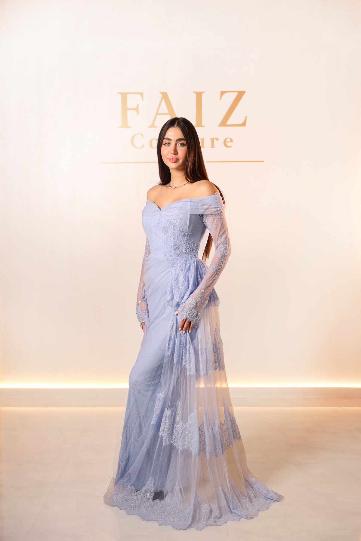 A woman modeling an off-the-shoulder light blue lace gown with long sheer sleeves and a fitted, tiered skirt, FAIZ Couture