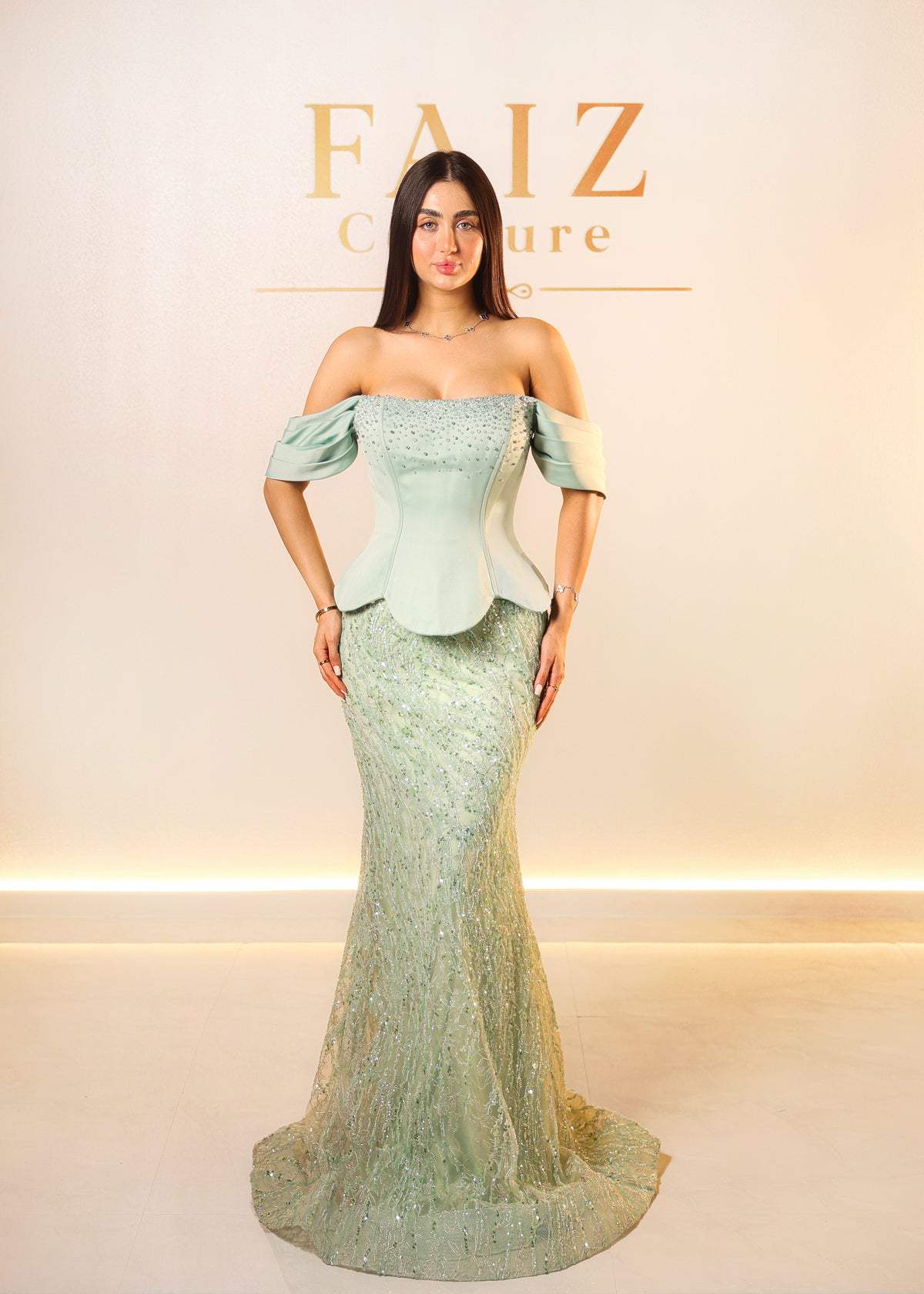 A woman modeling an off-the-shoulder mint green evening gown with a structured corset-style bodice and a flared embellished skirt, FAIZ Couture.