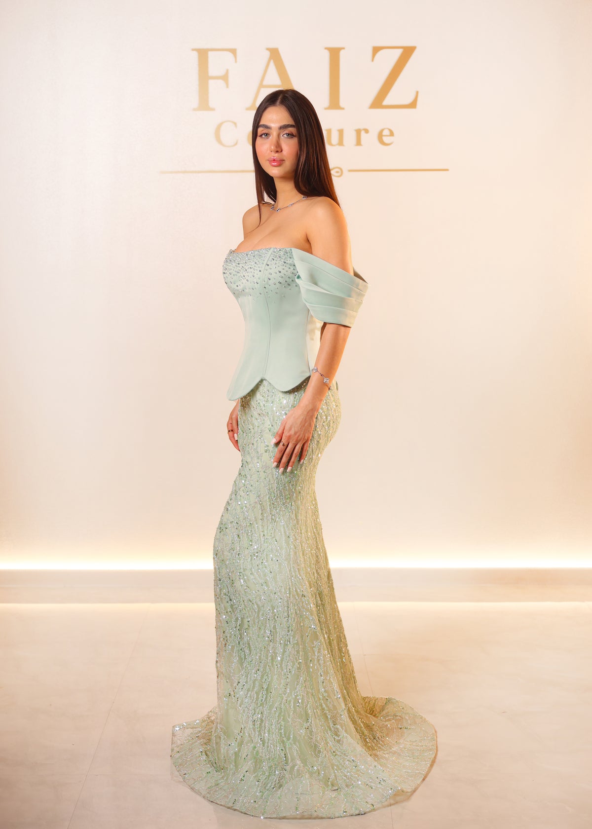 A woman models a light mint green off-the-shoulder evening gown with a structured corset-style bodice and a fitted lace mermaid skirt, FAIZ Couture.
