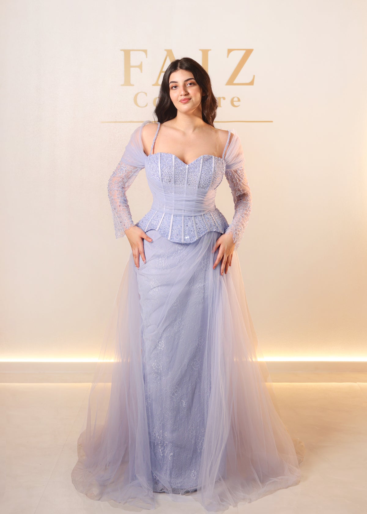 A woman modeling a light lavender blue formal gown with a structured corset bodice, sheer long sleeves, and a layered tulle skirt, FAIZ COUTURE
