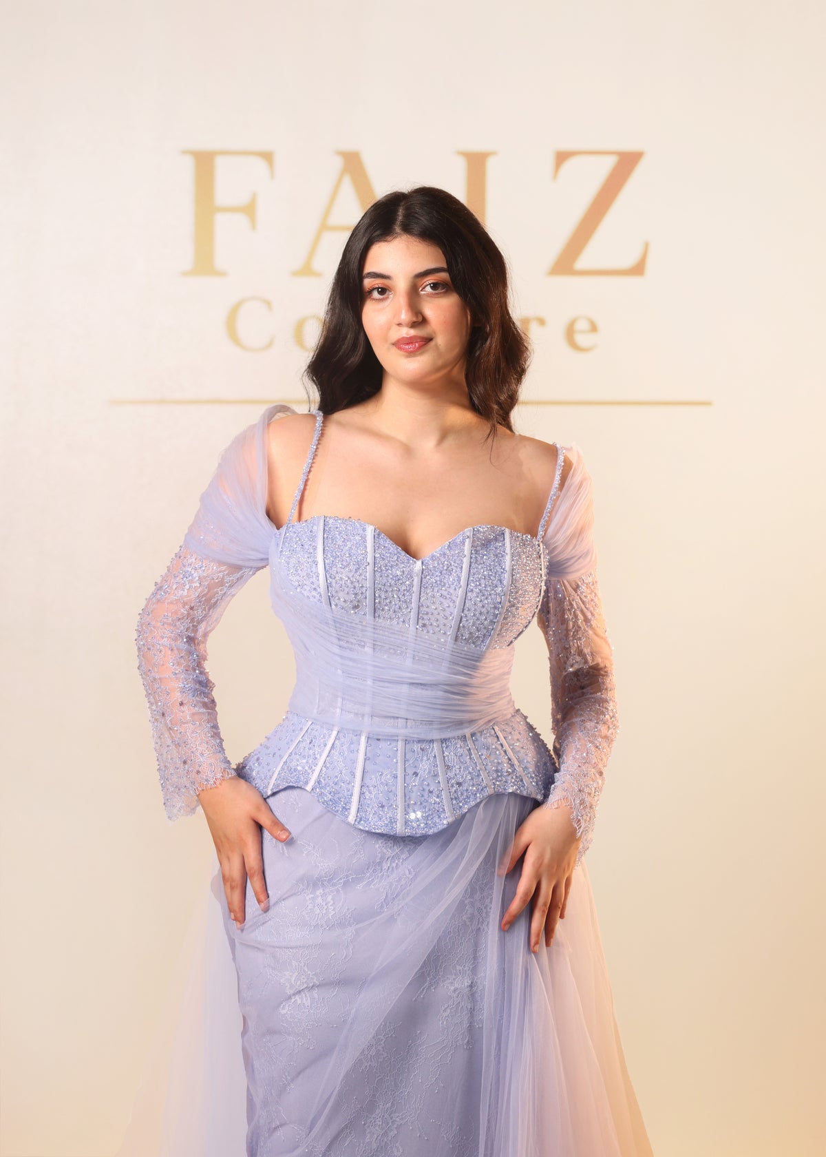 A woman modeling a light lavender blue gown with a structured corset-style bodice, sheer long sleeves, and a flowing skirt, FAIZ COUTURE