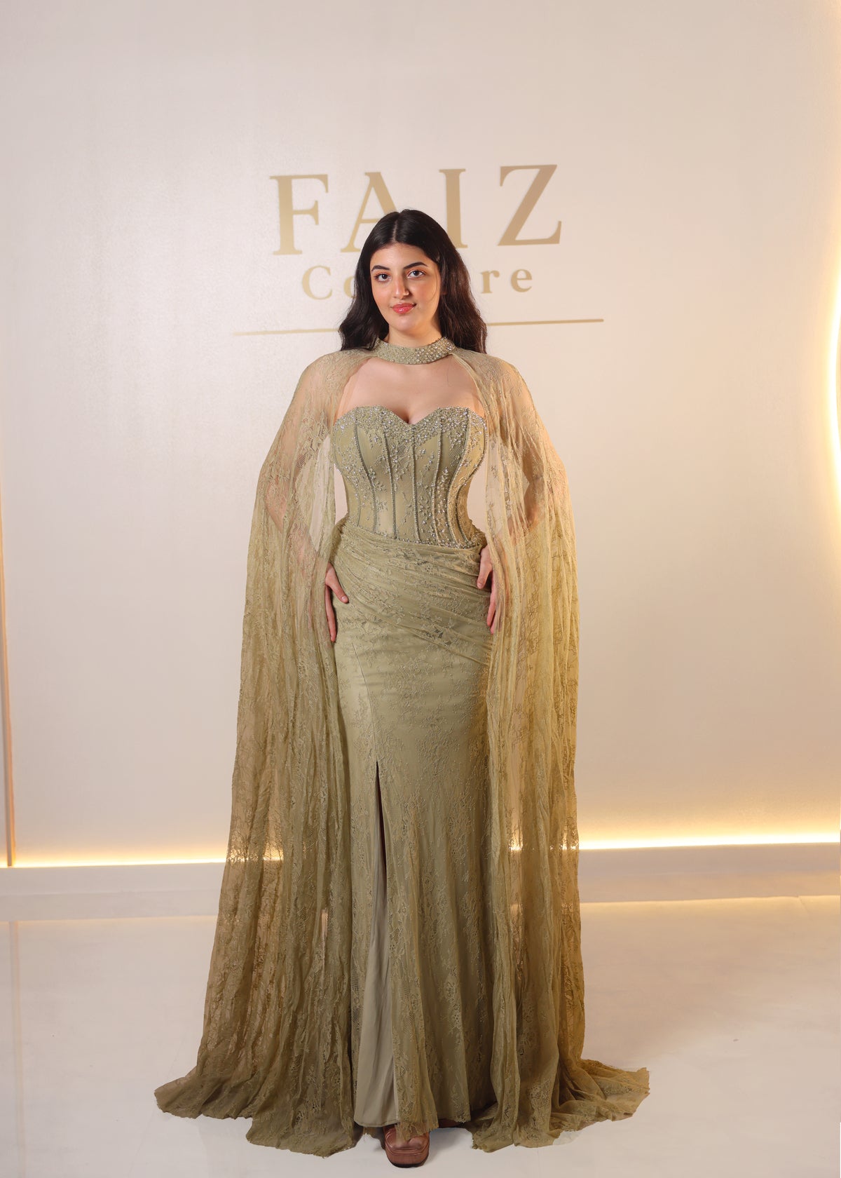 A woman stands in a studio wearing a floor-length olive green gown with a structured corset-style bodice, sheer draped cape sleeves, and a high slit. FAIZ COUTURE