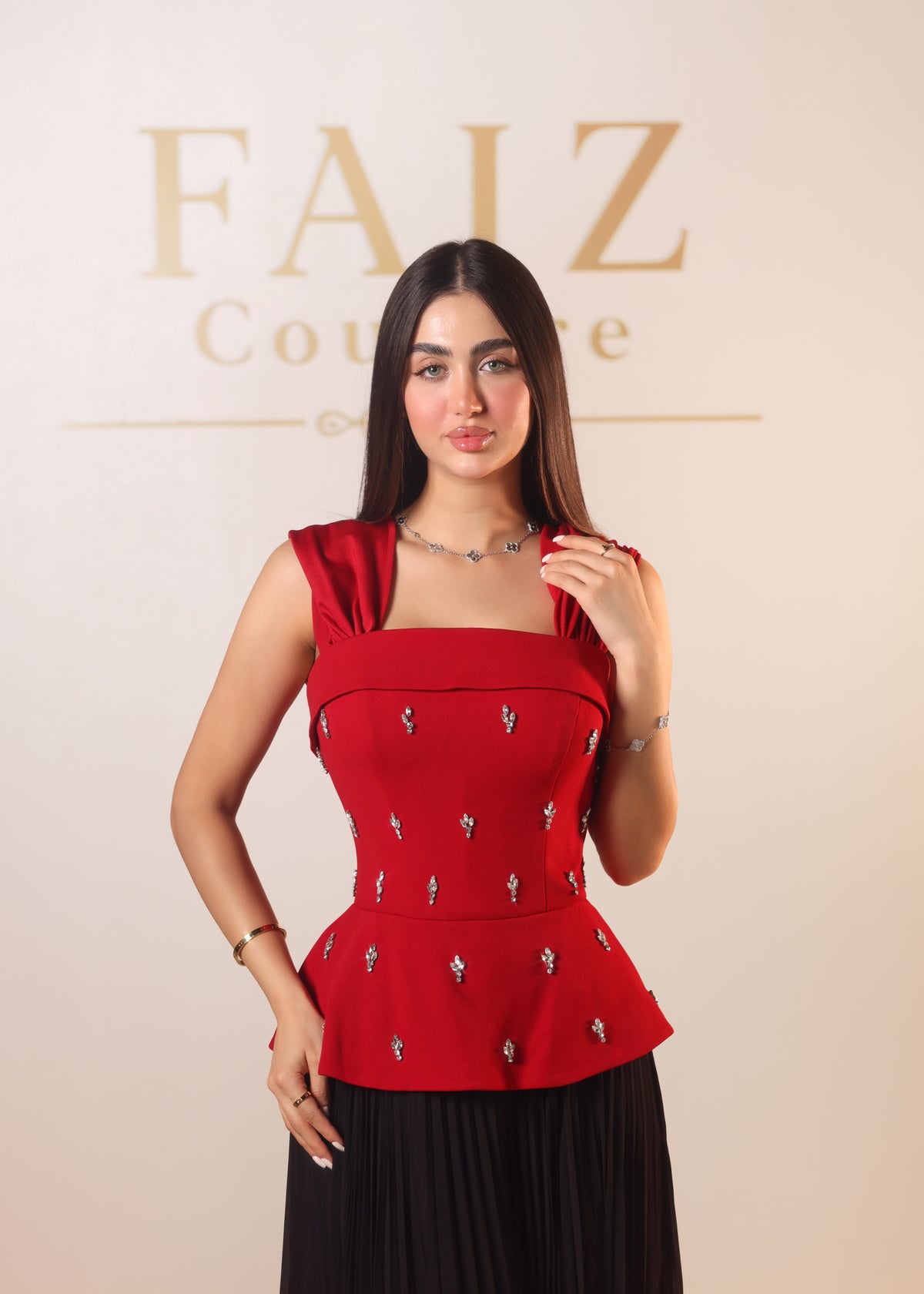 A woman modeling a red sleeveless top with a square neckline and silver embellishments, paired with a black pleated skirt, FAIZ COUTURE