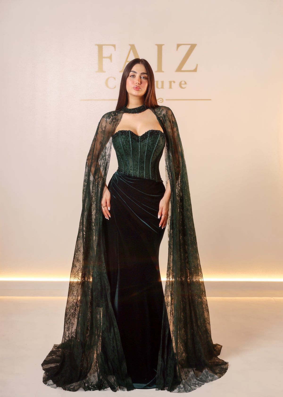 A woman models a dark green and black evening gown with a sheer lace cape and structured corset bodice,  FAIZ COUTURE