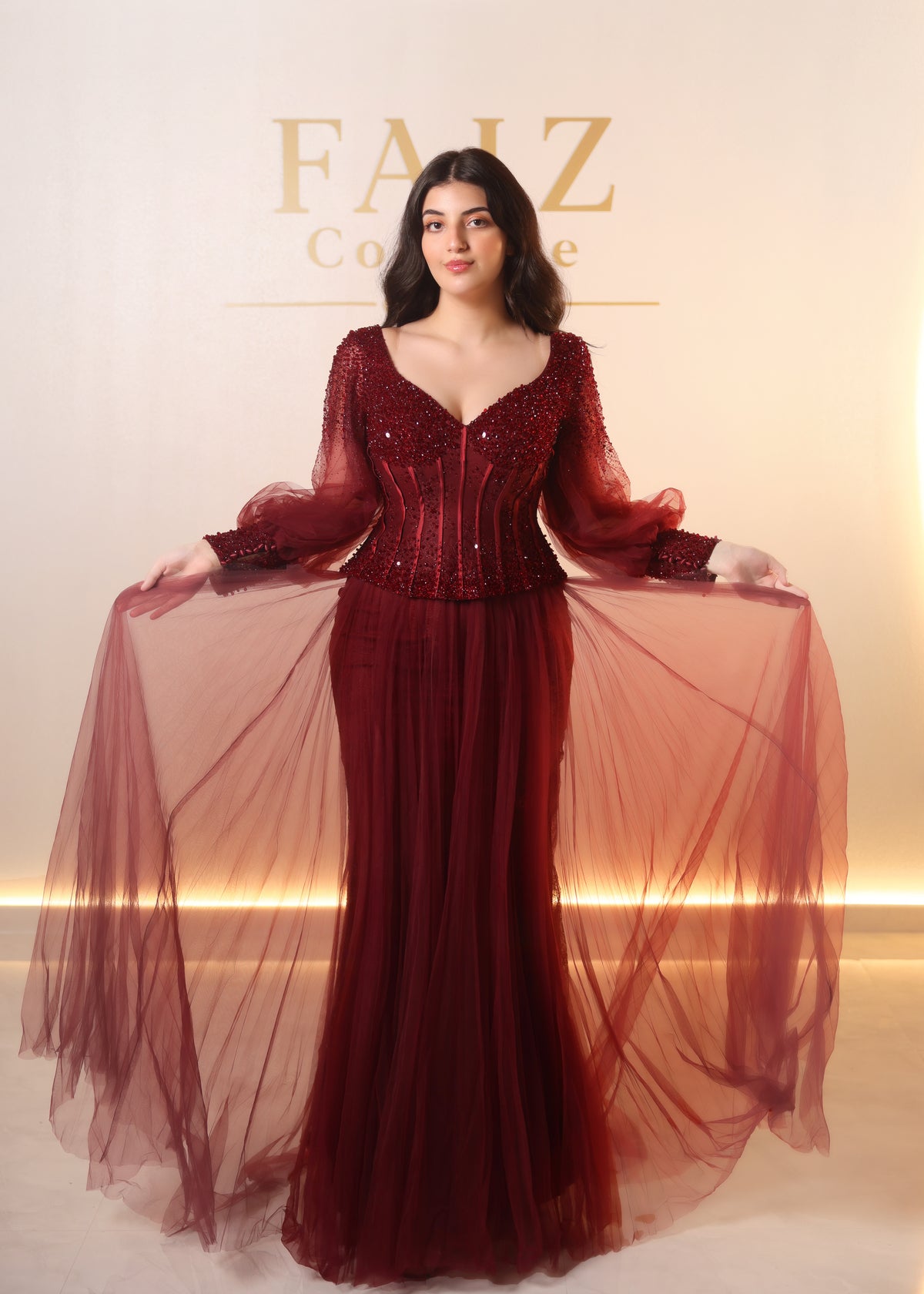 A woman modeling a deep burgundy red evening gown with sheer long sleeves, a beaded corset-style bodice, and a voluminous tulle skirt, Faiz Couture