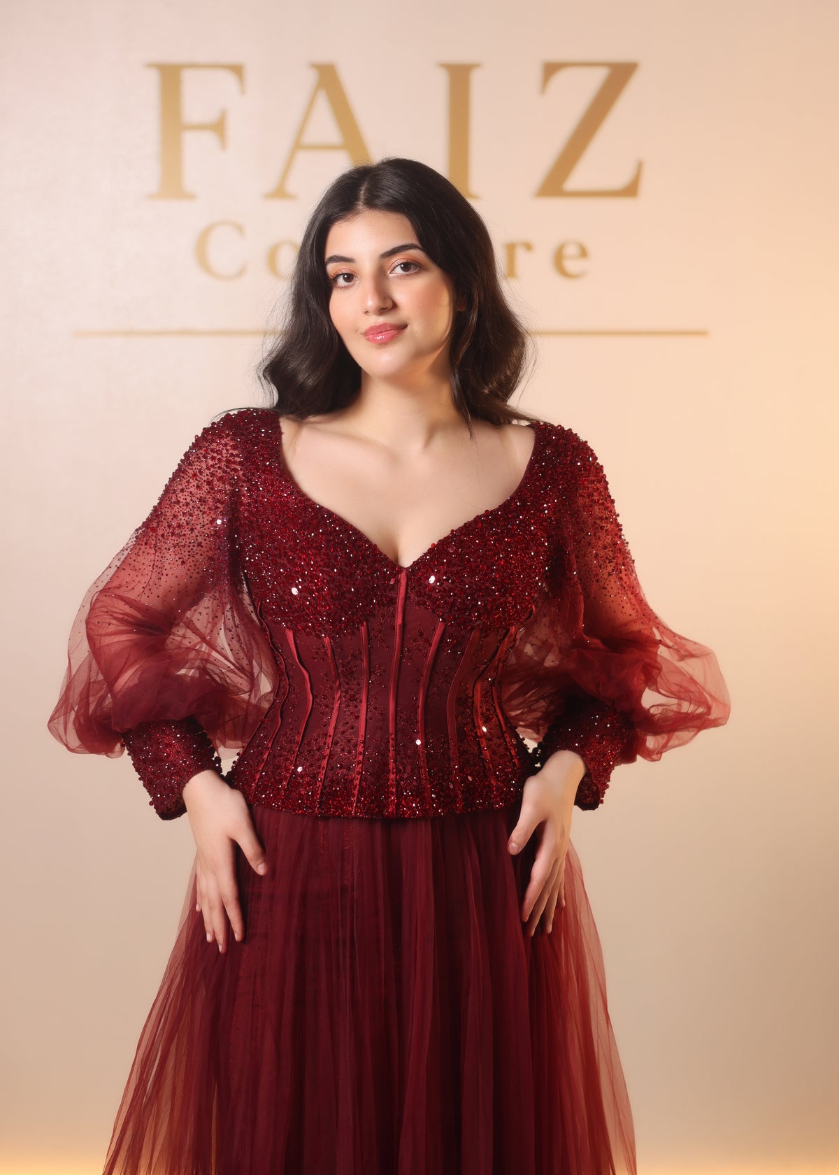 A woman modeling a deep burgundy red evening gown with a heavily embellished sequined corset bodice and sheer puff sleeves, Faiz Couture