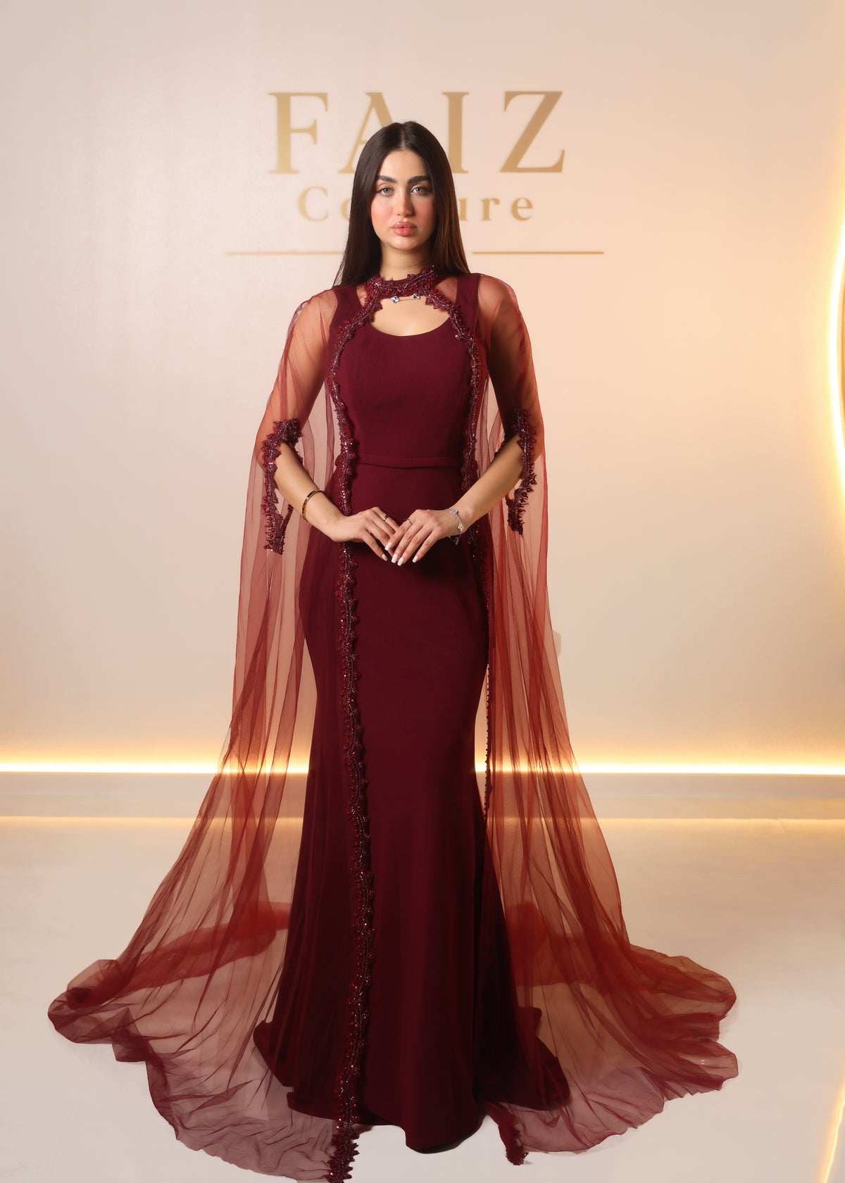 A woman modeling a floor-length burgundy gown with a sheer draped cape overlay and dark floral embroidery, FAIZ COUTURE