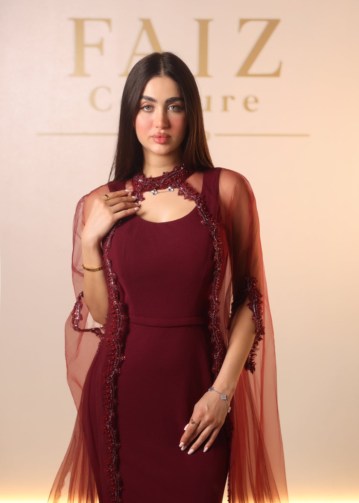 A woman modeling a sleeveless deep burgundy dress with sheer draped overlays and dark embroidered detailing, FAIZ COUTURE