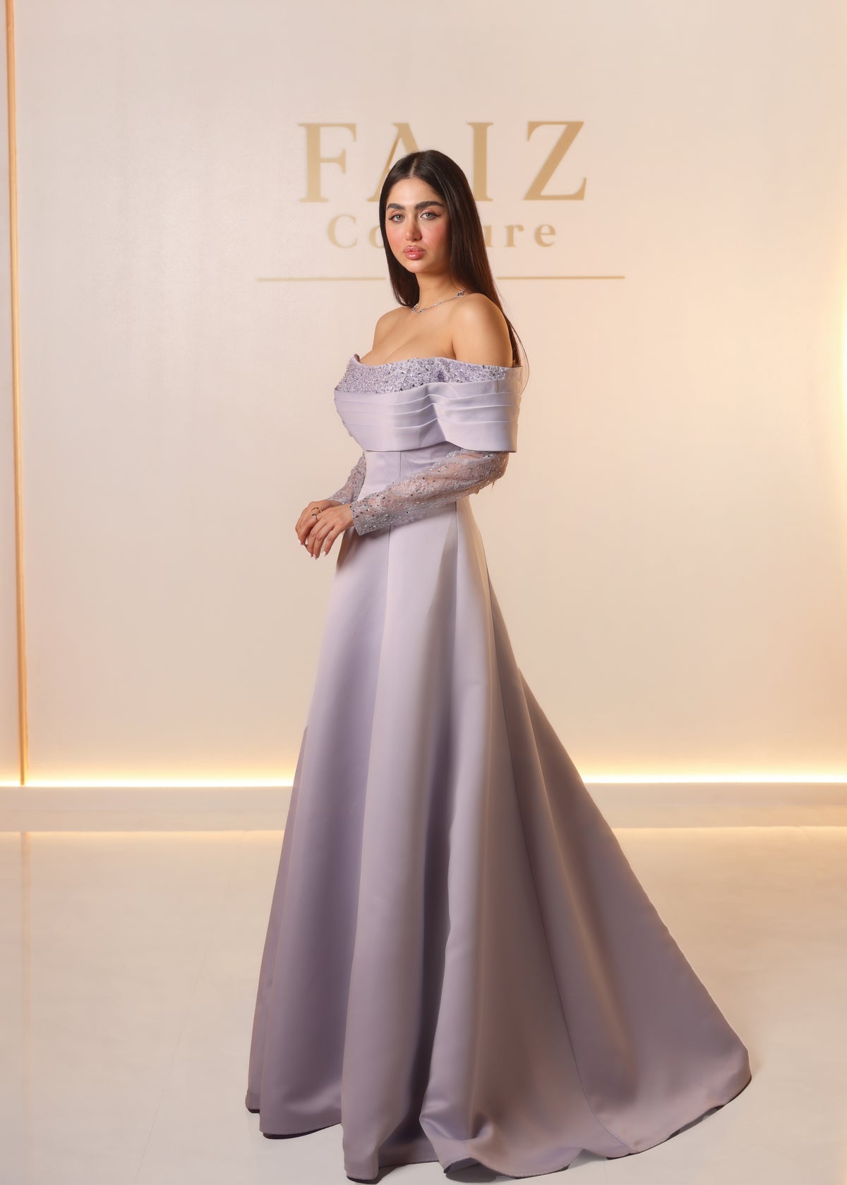 A woman modeling an off-the-shoulder floor-length gown in a soft lavender-gray satin fabric, FAIZ COUTURE