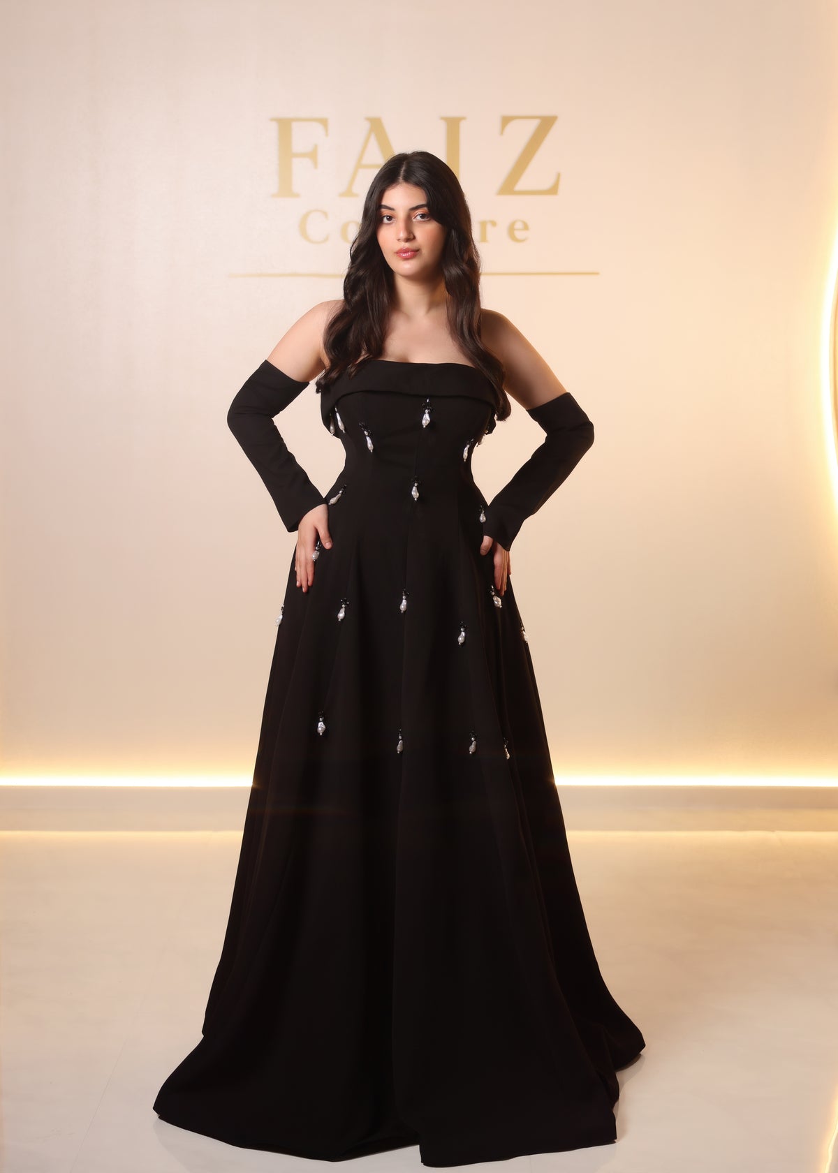 A woman modeling a strapless black floor-length gown with long sleeves and small silver embellishments, Faiz Couture