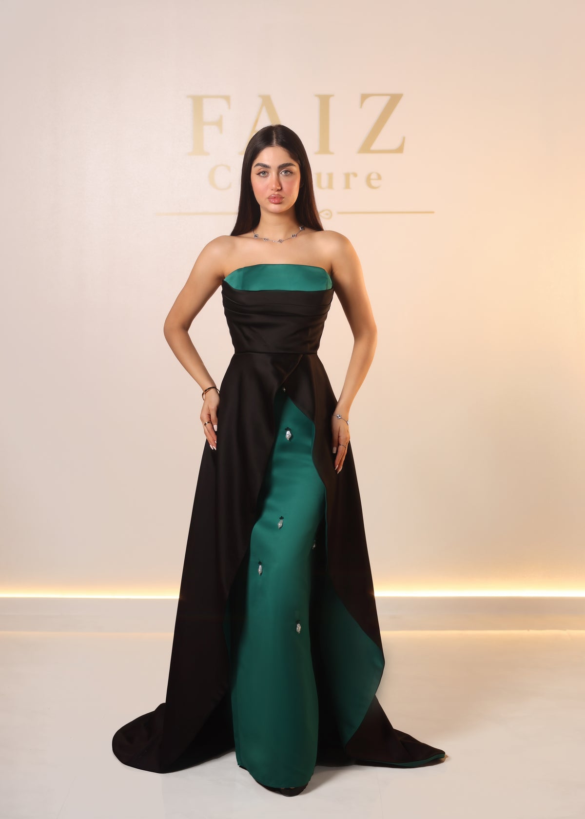A woman models a strapless high-low evening gown with a black outer layer and a teal green inner skirt. FAIZ COUTURE