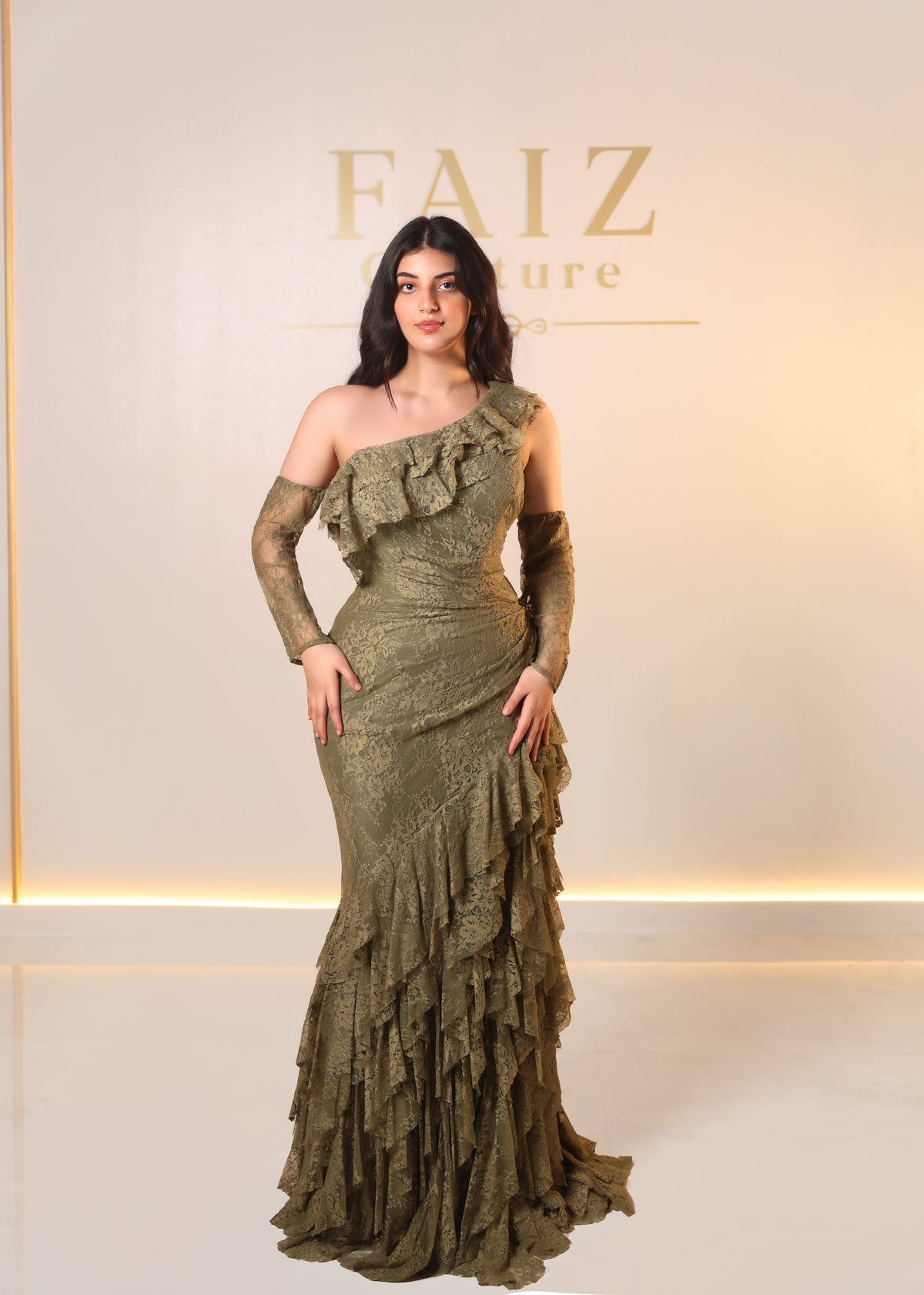 A woman models a floor-length olive green ruffled gown with long sheer sleeves and a structured one-shoulder neckline, FAIZ COUTURE