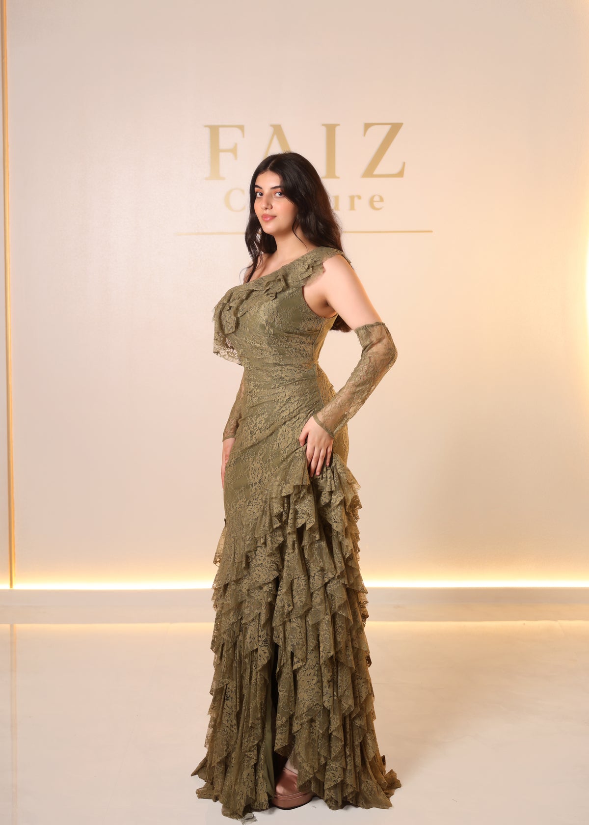 A woman models a floor-length olive green ruffled gown with long sheer sleeves and a one-shoulder neckline, FAIZ COUTURE