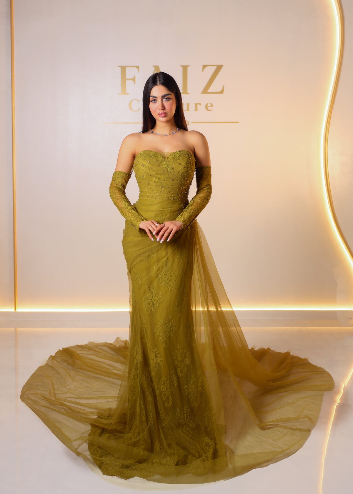 A woman models a strapless olive green lace gown with long sleeves and a sheer tulle train, FAIZ COUTURE