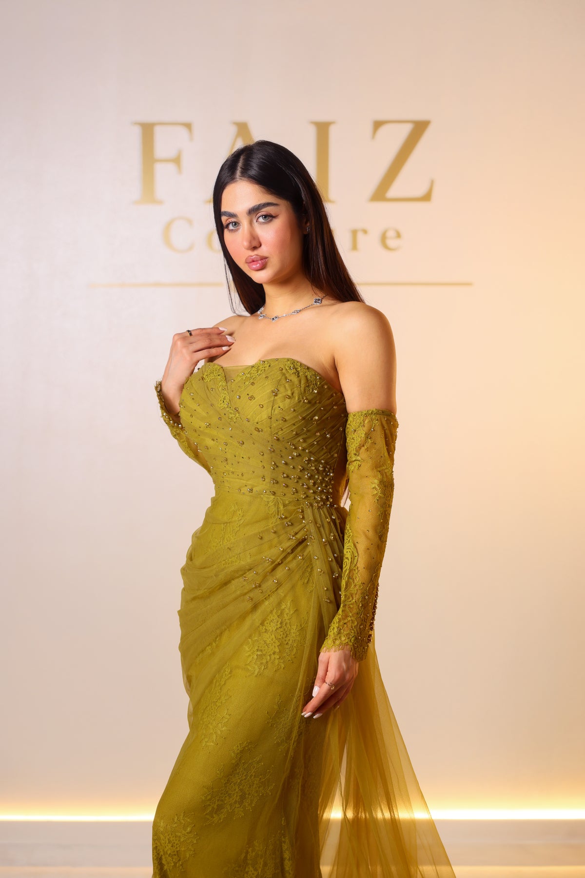 A woman modeling a strapless olive green gown with long lace sleeves and a sheer tulle overlay, FAIZ COUTURE