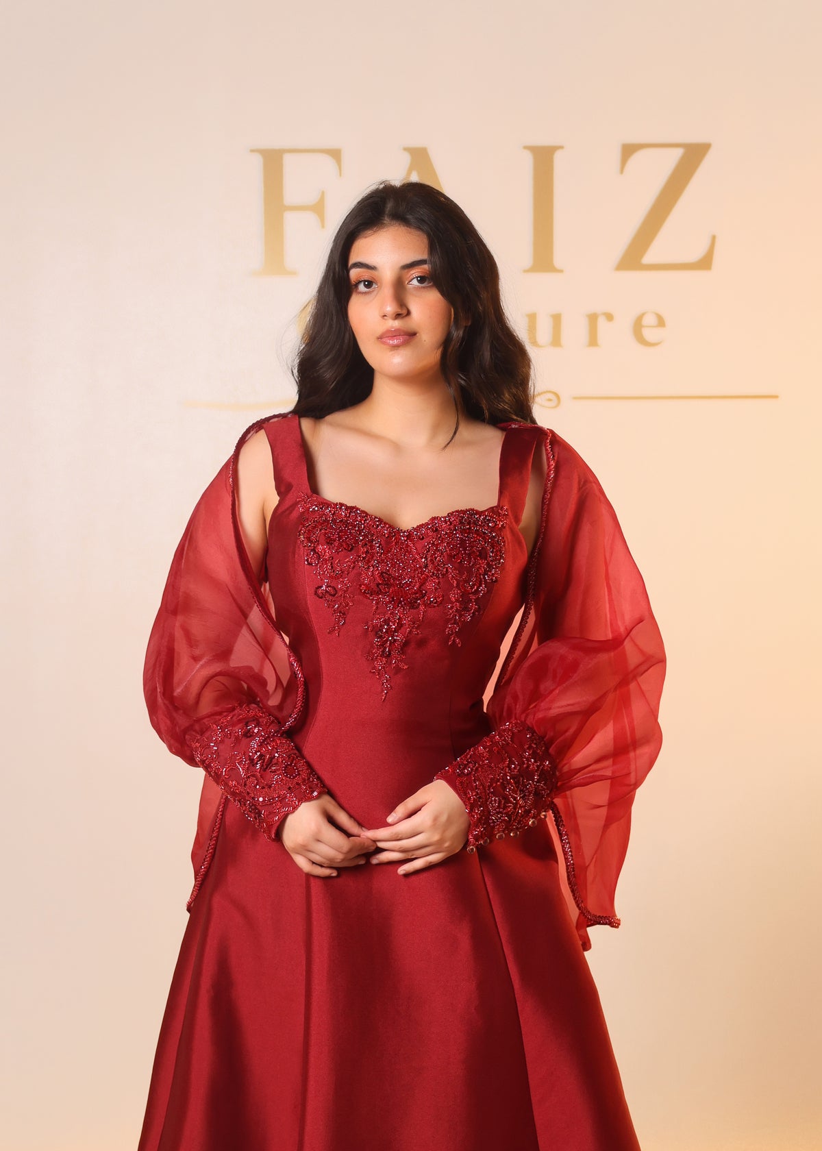 A woman wearing a deep red evening gown with sheer draped sleeves and intricate floral embroidery on the bodice, FAIZ COUTURE