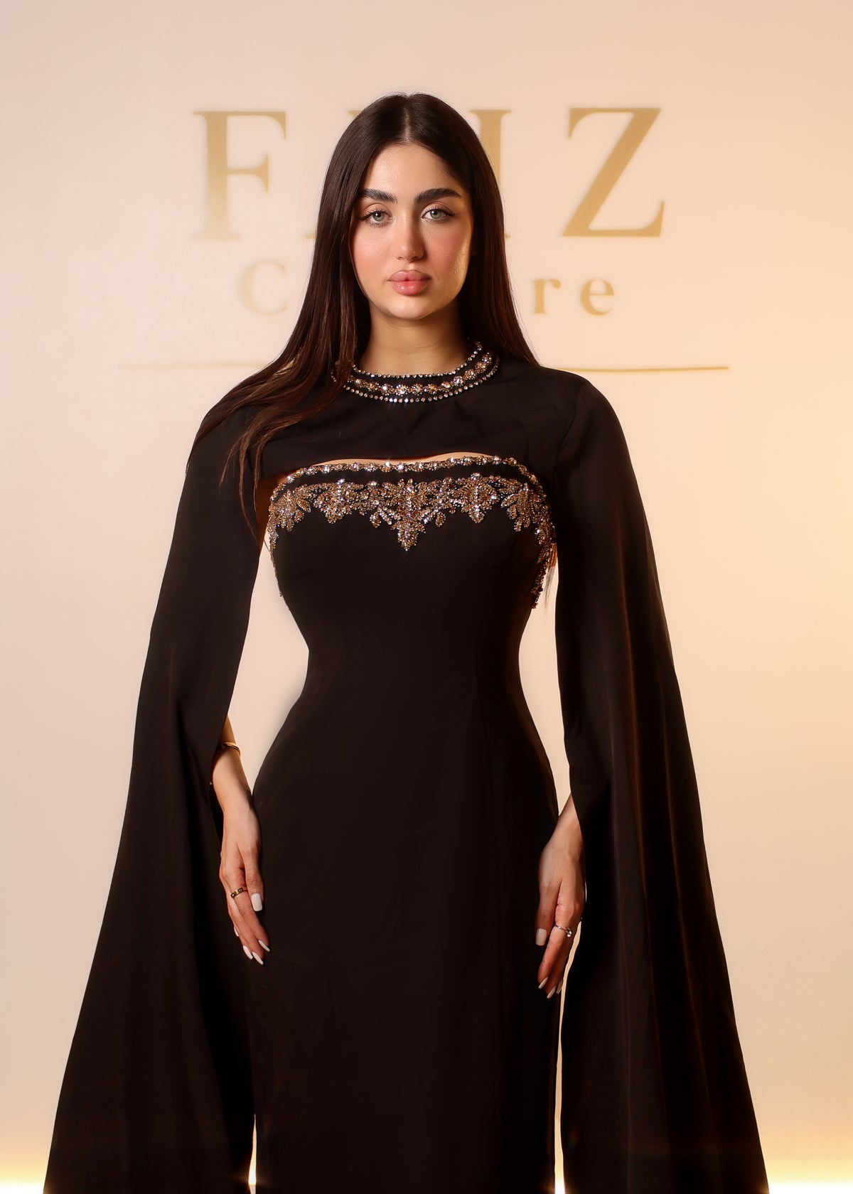 A woman modeling a black floor-length dress with a cape-like draped sleeve and ornate gold embroidery across the chest and neckline, FAIZ COUTURE