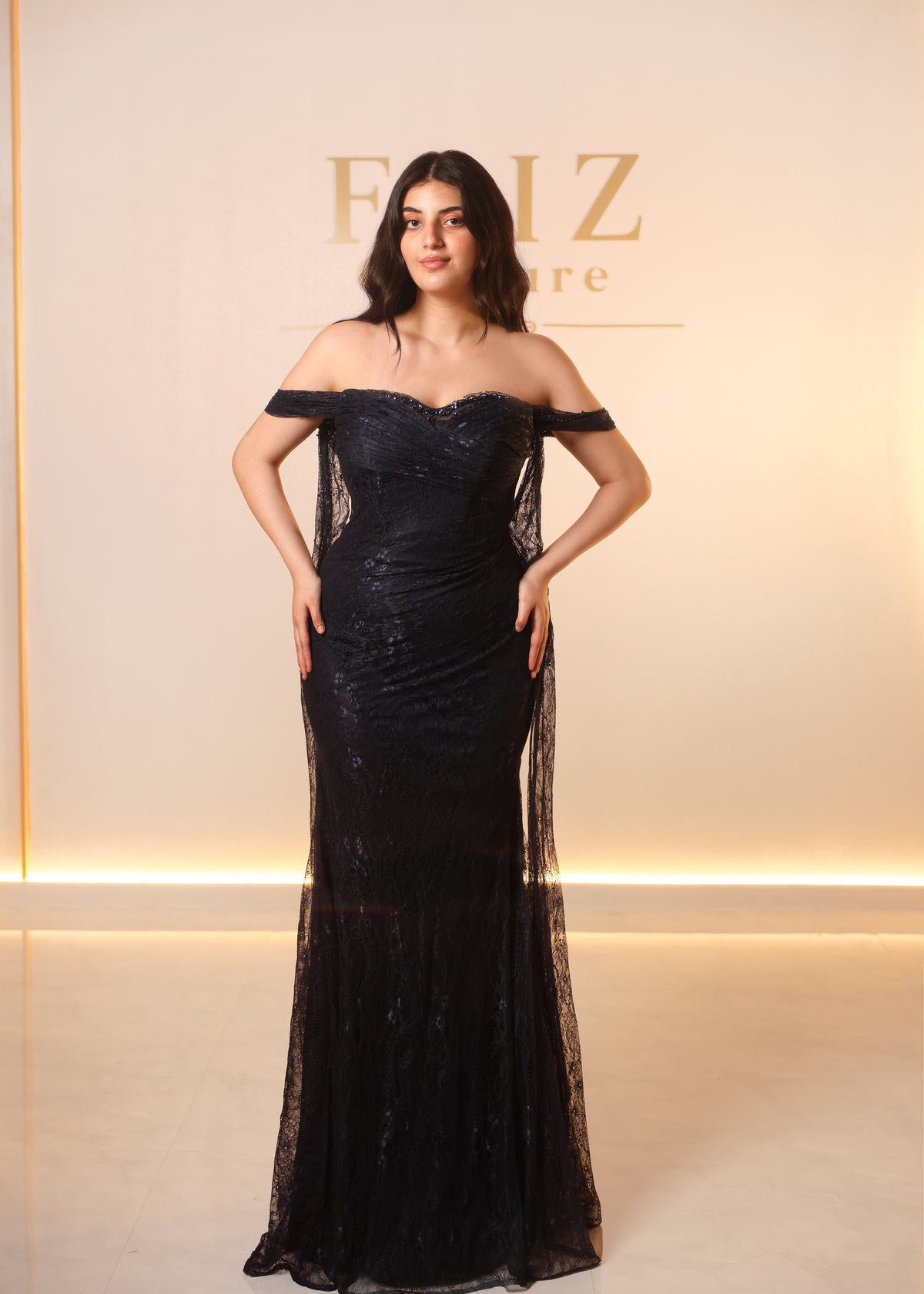 A woman modeling an off-the-shoulder black lace evening gown with a fitted bodice and sheer side panels, FAIZ COUTURE