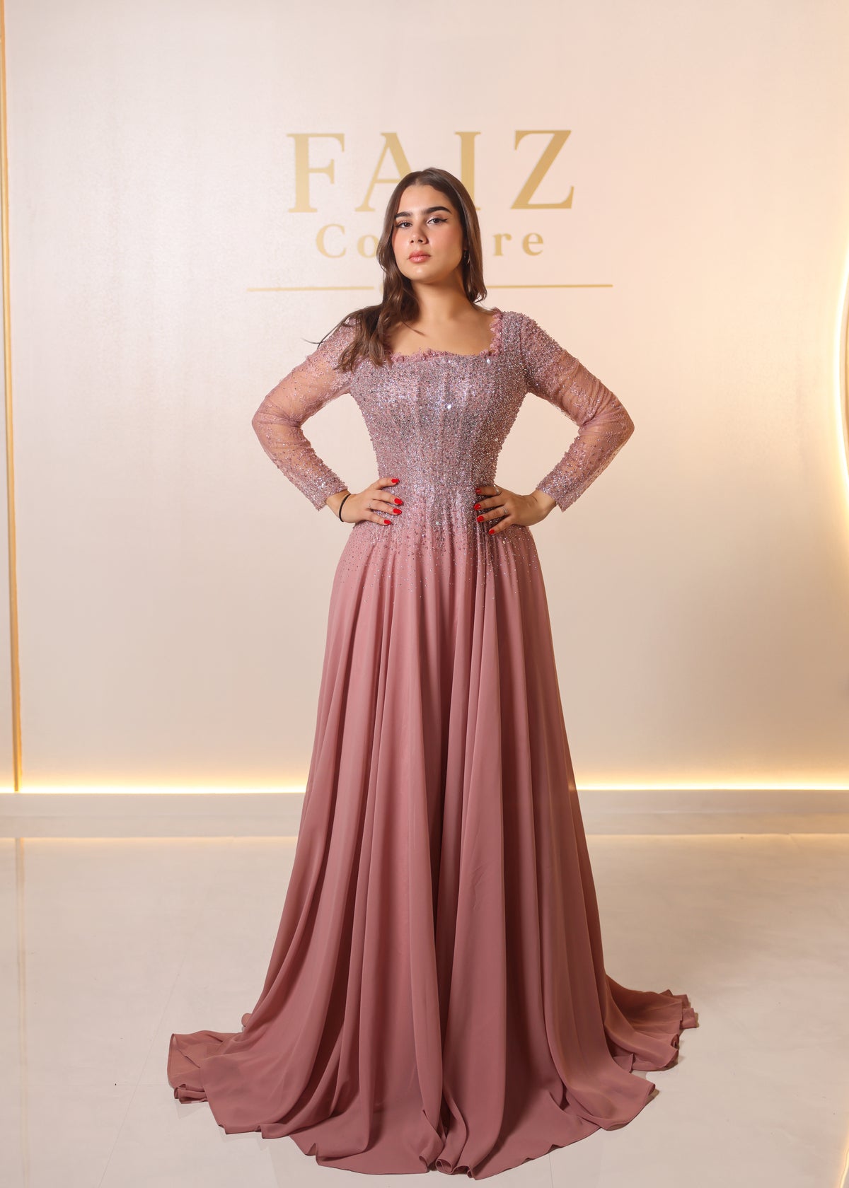 A woman modeling a floor-length dusty rose formal gown with a beaded lace bodice and long sheer sleeves. FAIZ COUTURE