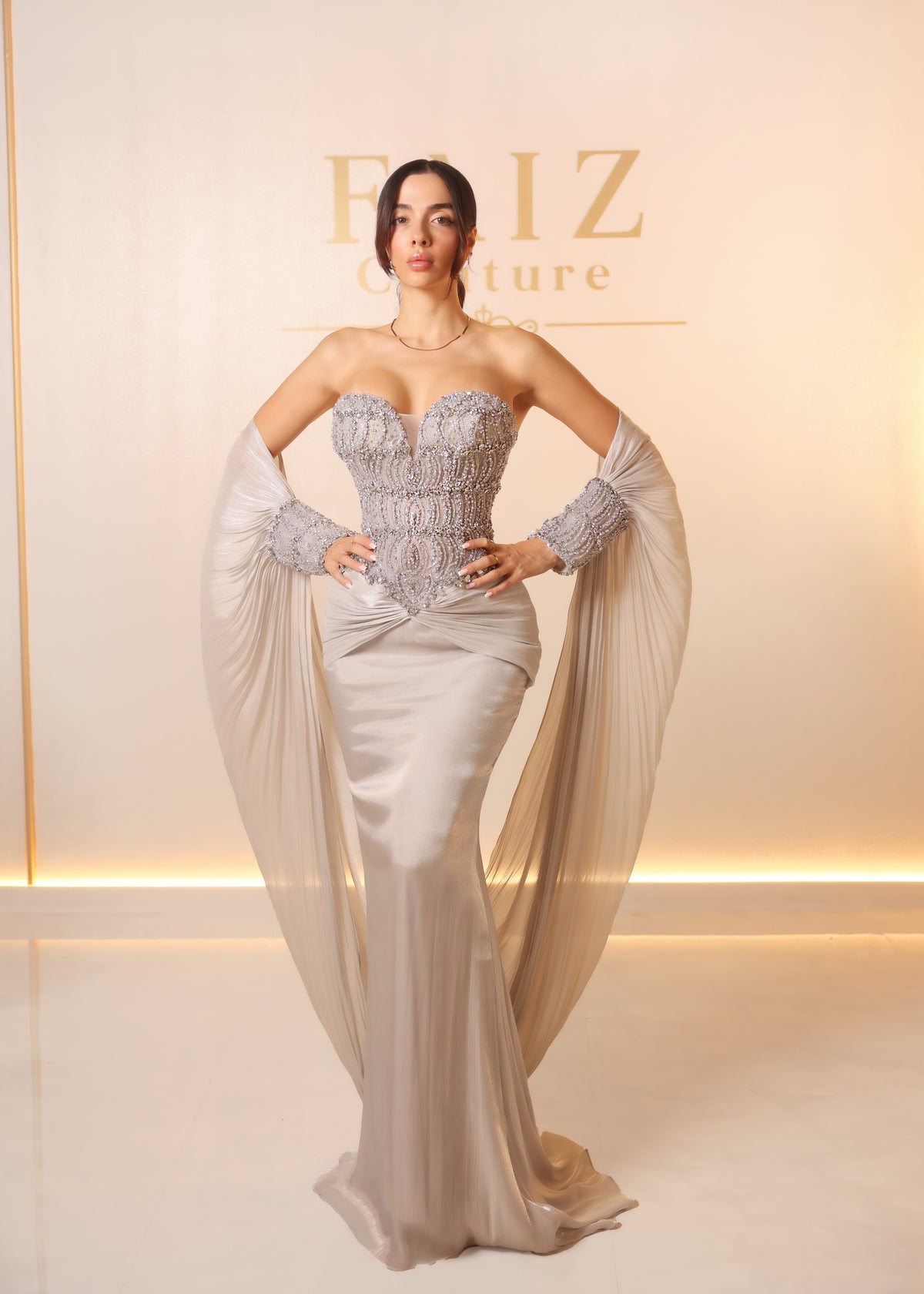 A woman models a strapless champagne beige evening gown with a heavily embellished silver lace corset bodice, matching lace gloves, and a long flowing chiffon overskirt. FAIZ COUTURE