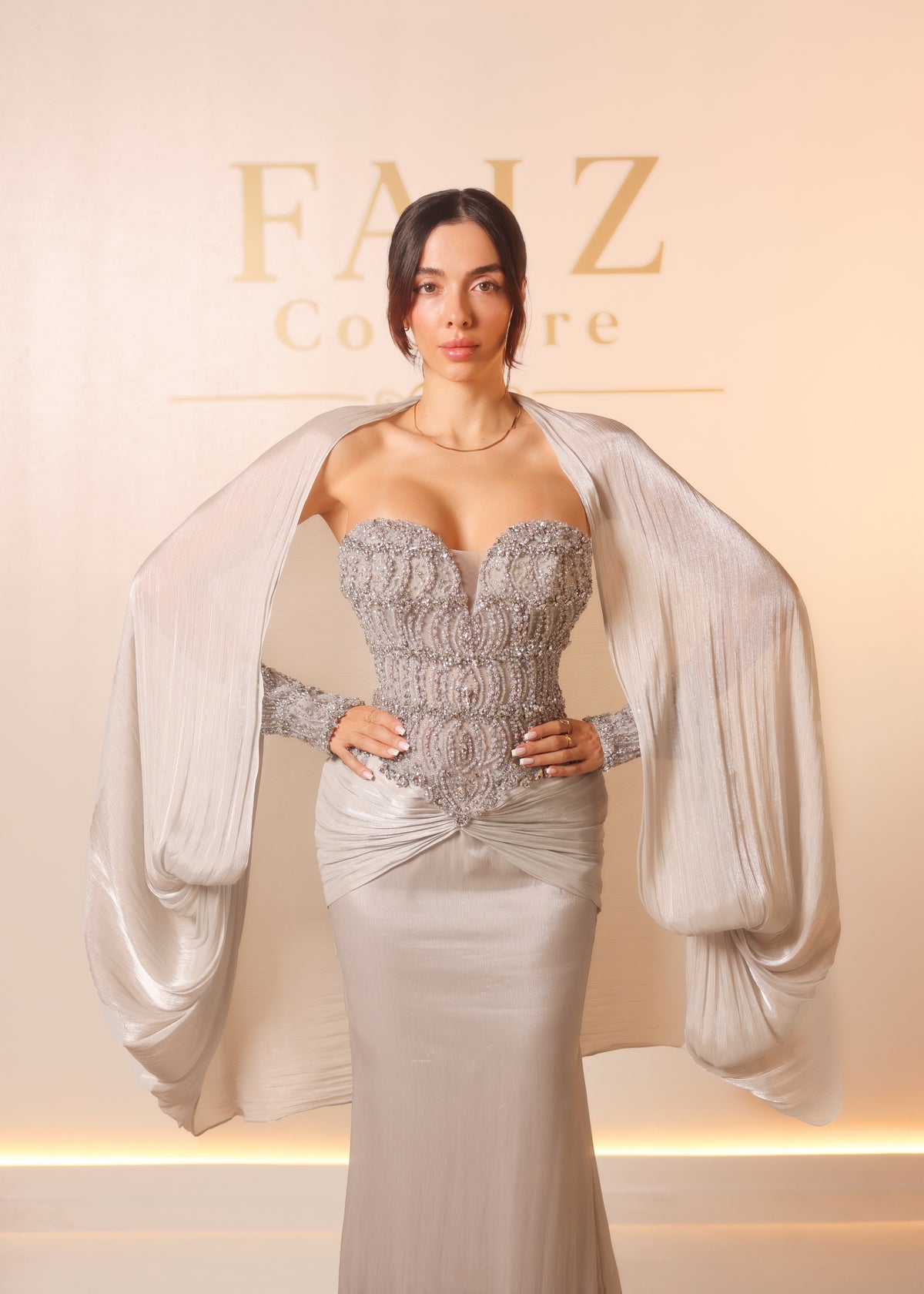 A woman models a strapless silver lace corset dress with a matching sheer draped cape, FAIZ COUTURE