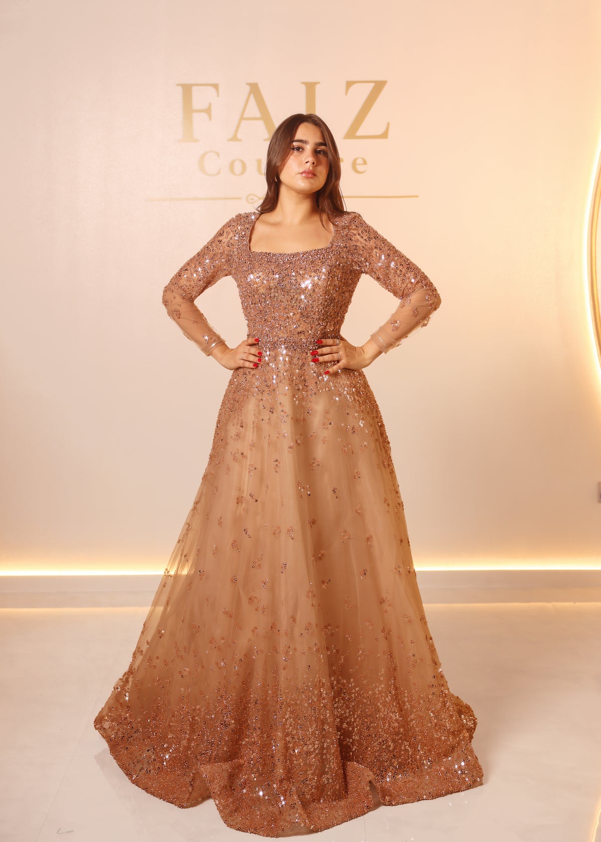 A woman modeling a floor-length, long-sleeve evening gown with heavy sequin and bead embellishment in a warm champagne-gold tone. FAIZ COUTURE