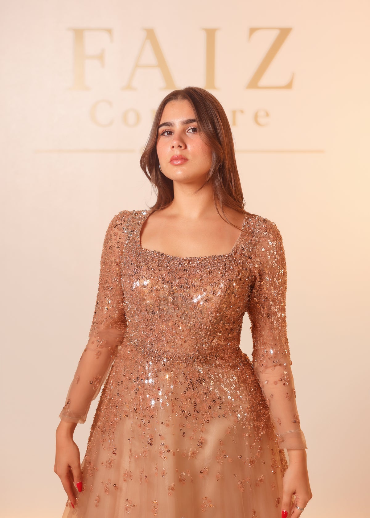 A woman modeling a sparkling champagne sequined gown with sheer long sleeves and a square neckline. FAIZ COUTURE