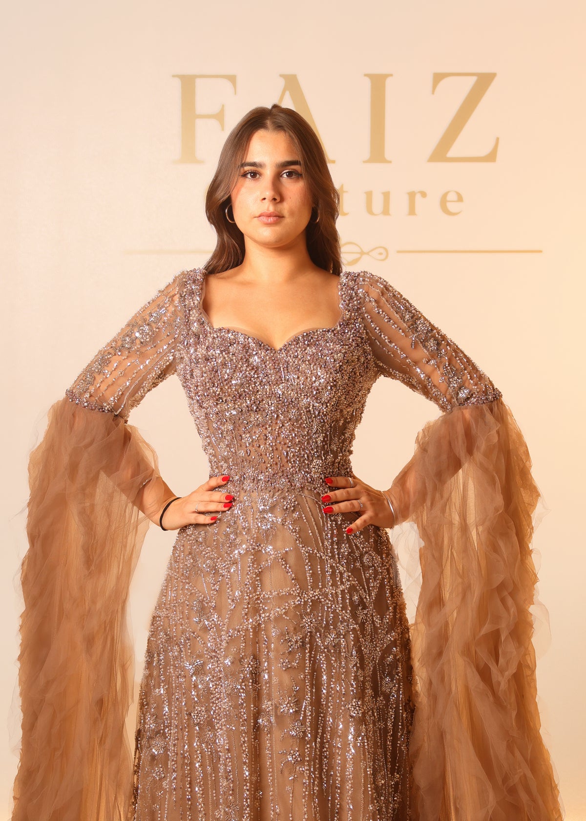 A woman wearing a heavily embellished champagne beige gown with sheer long sleeves and dramatic tulle extensions, FAIZ couture
