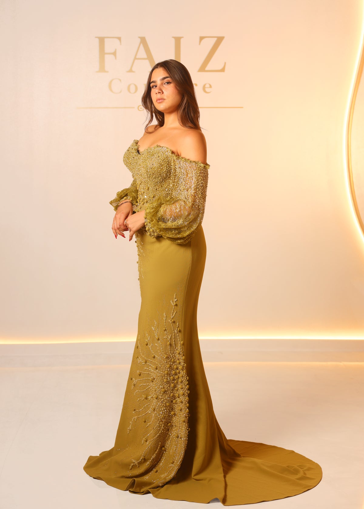A woman modeling an off-the-shoulder olive green evening gown with intricate lace and bead embellishments, FAIZ COUTURE