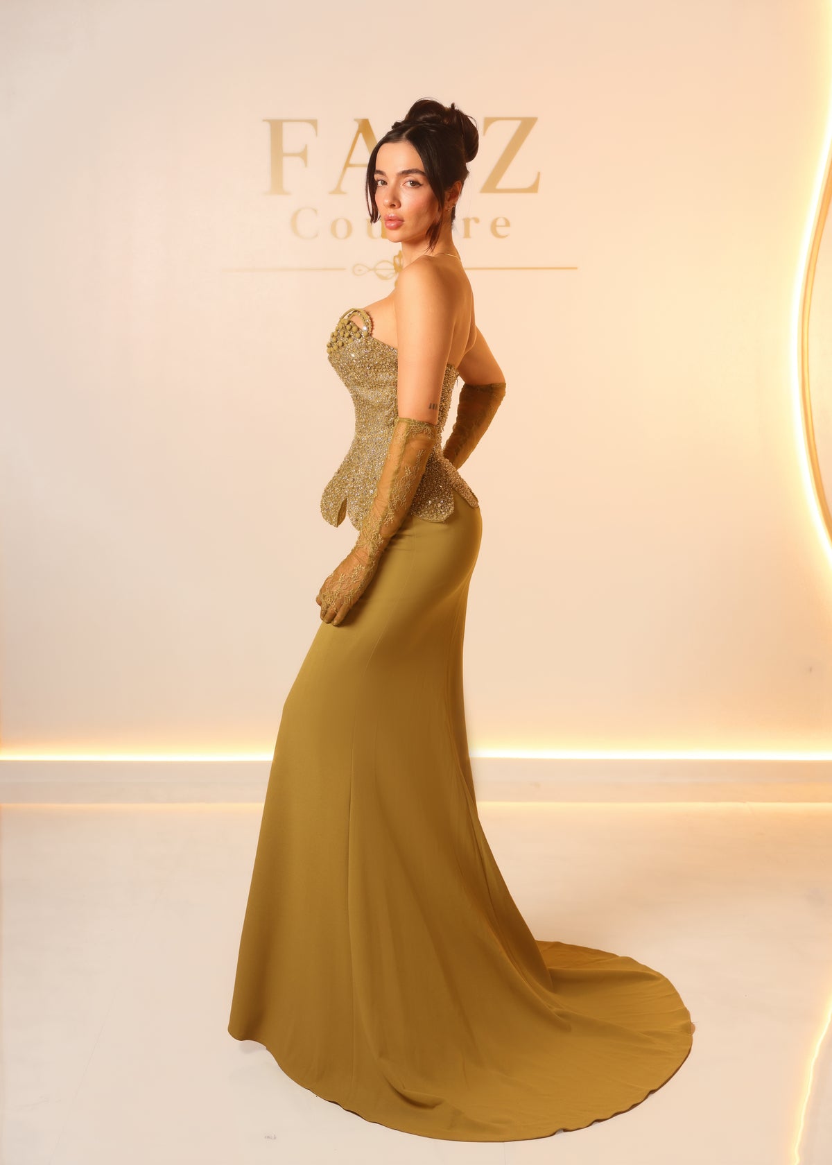 A woman modeling a floor-length evening gown in a warm olive-gold color, featuring a structured corset-style bodice with metallic gold lace applique and matching lace gloves. FAIZCOUTURE