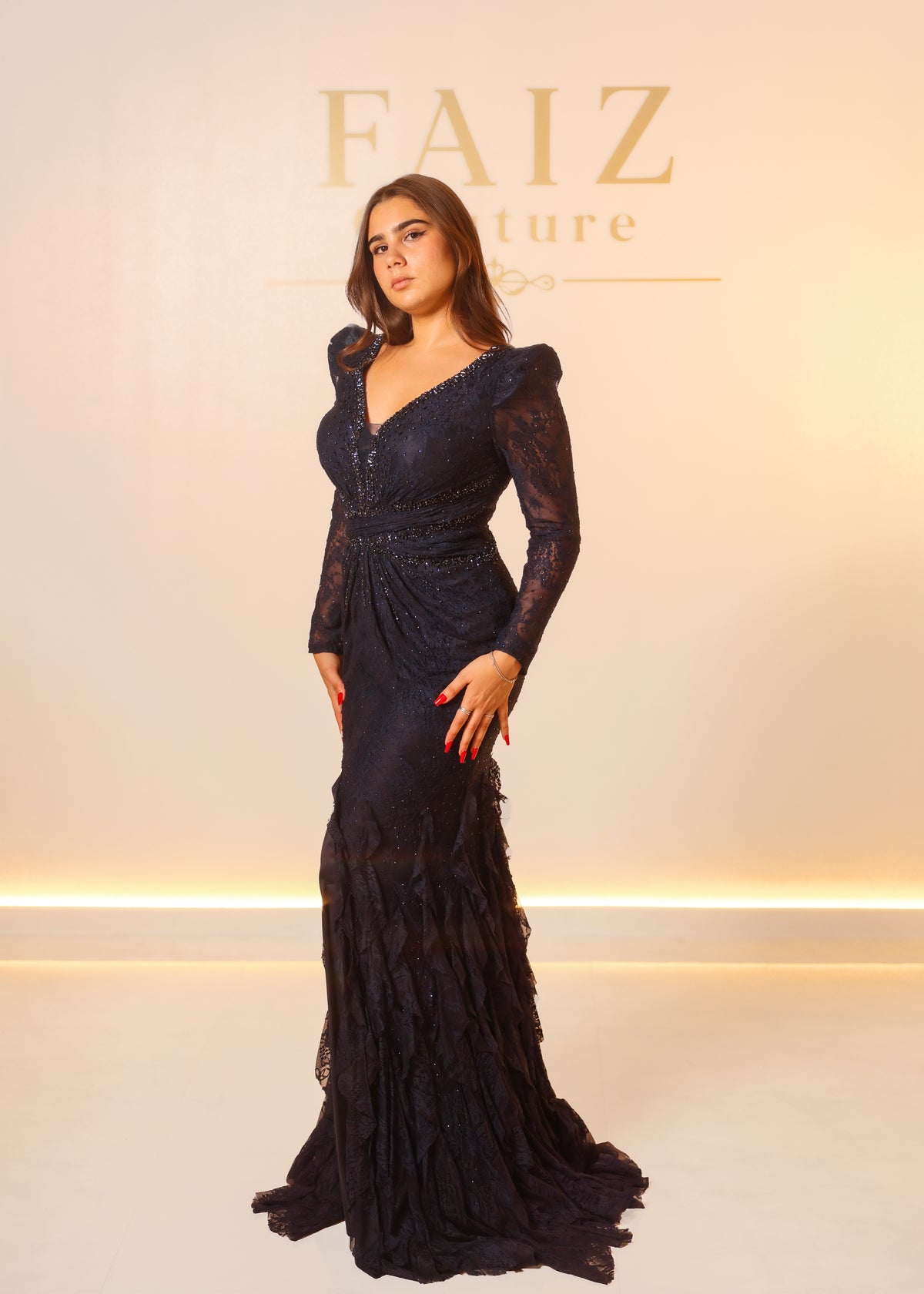 A woman modeling a long-sleeved, floor-length navy lace gown with a fitted silhouette and ruffled hem, FAIZ couture