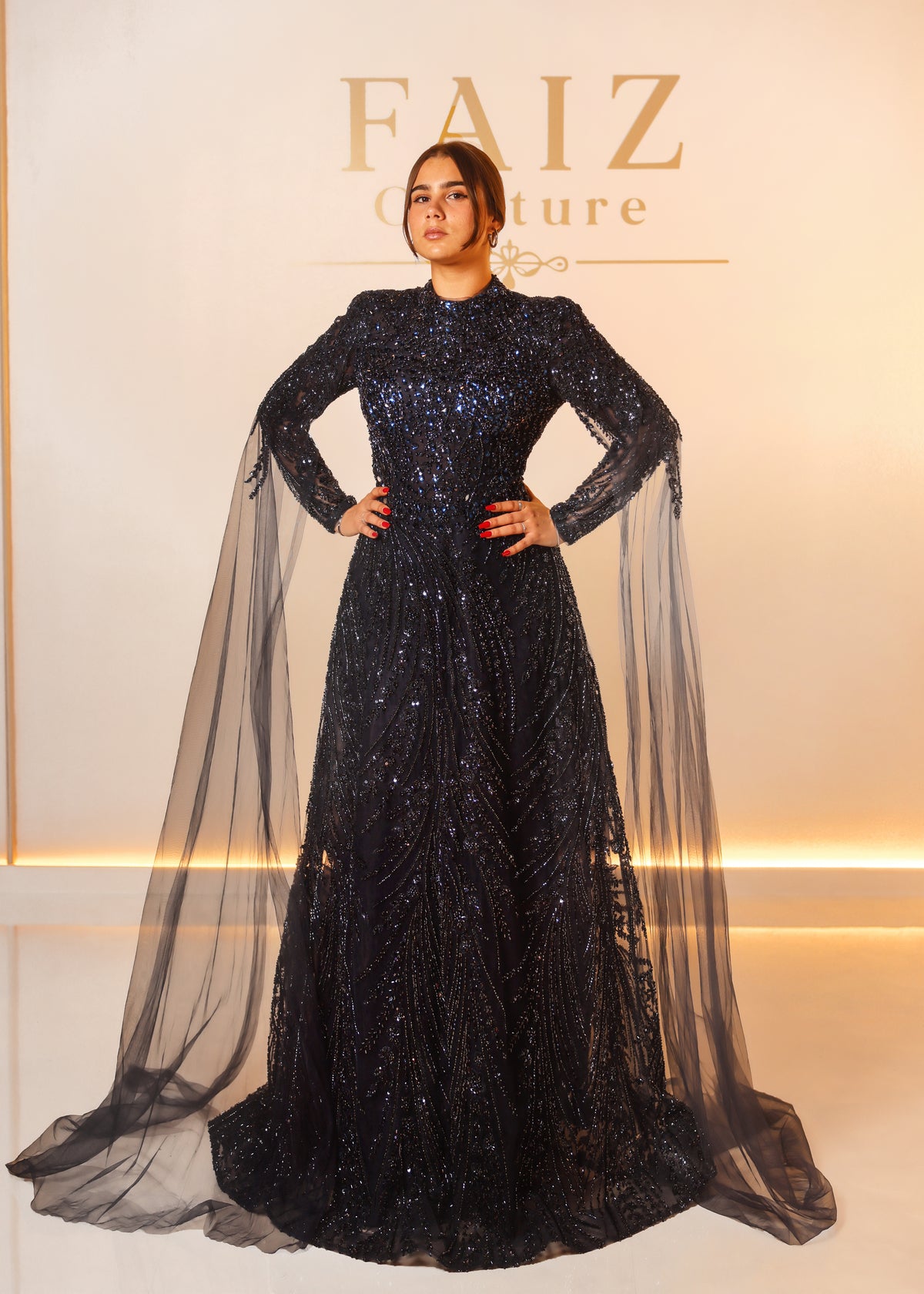 A woman modeling a long-sleeved, floor-length black gown covered in shimmering sequins, featuring sheer draped cape sleeves, FAIZ COUTURE