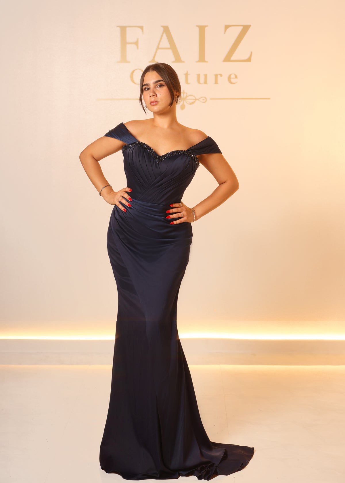A woman modeling a floor-length off-the-shoulder navy gown with a fitted bodice and slight train, FANIZ Couture