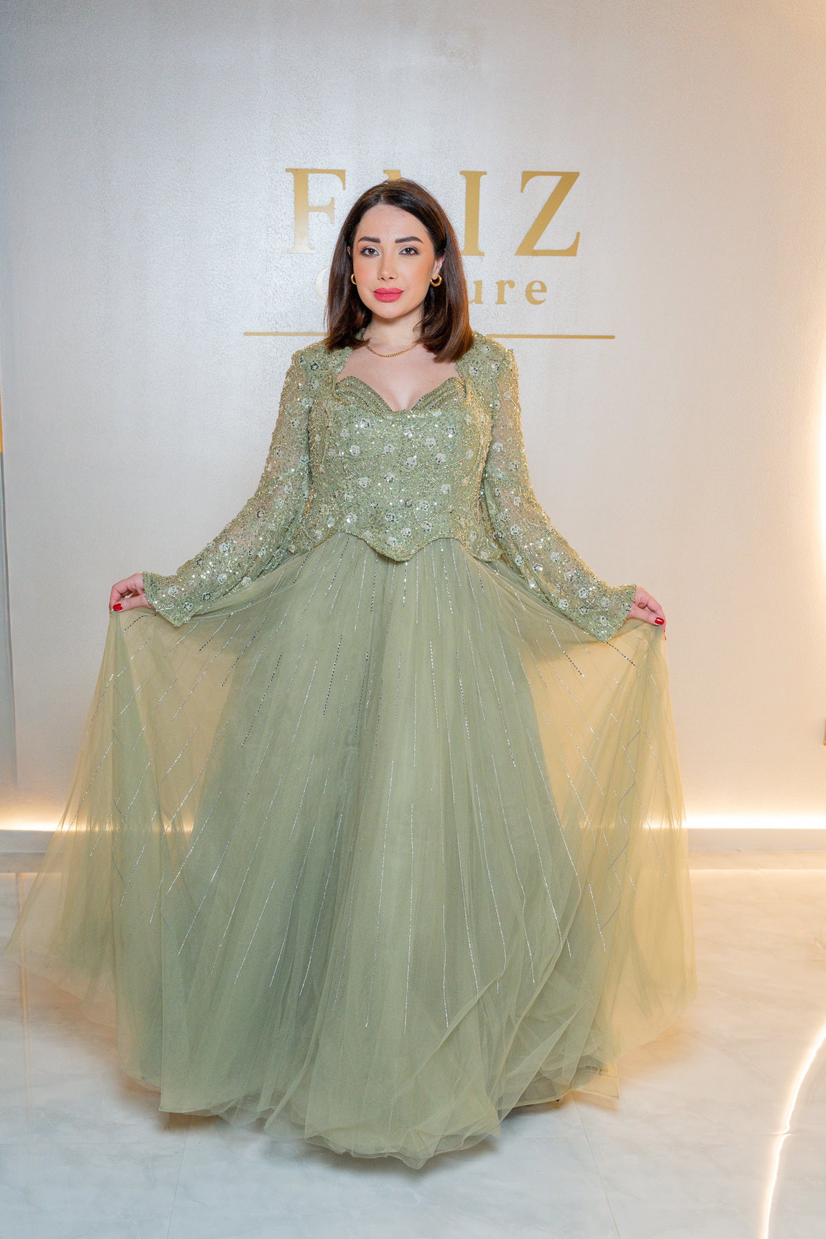 A woman modeling a floor-length sage green gown with a heavily embellished beaded bodice and long sheer sleeves, holding out the voluminous tulle skirt FAIZ COUTURE