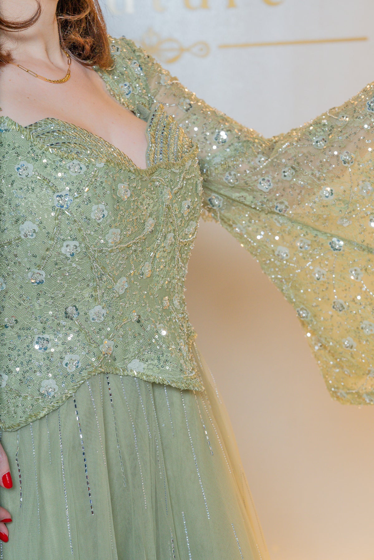 A close-up of a model wearing a light sage green embellished gown with a sweetheart neckline, heavily decorated with sequins and crystals, and dramatic sheer bell sleeves.
