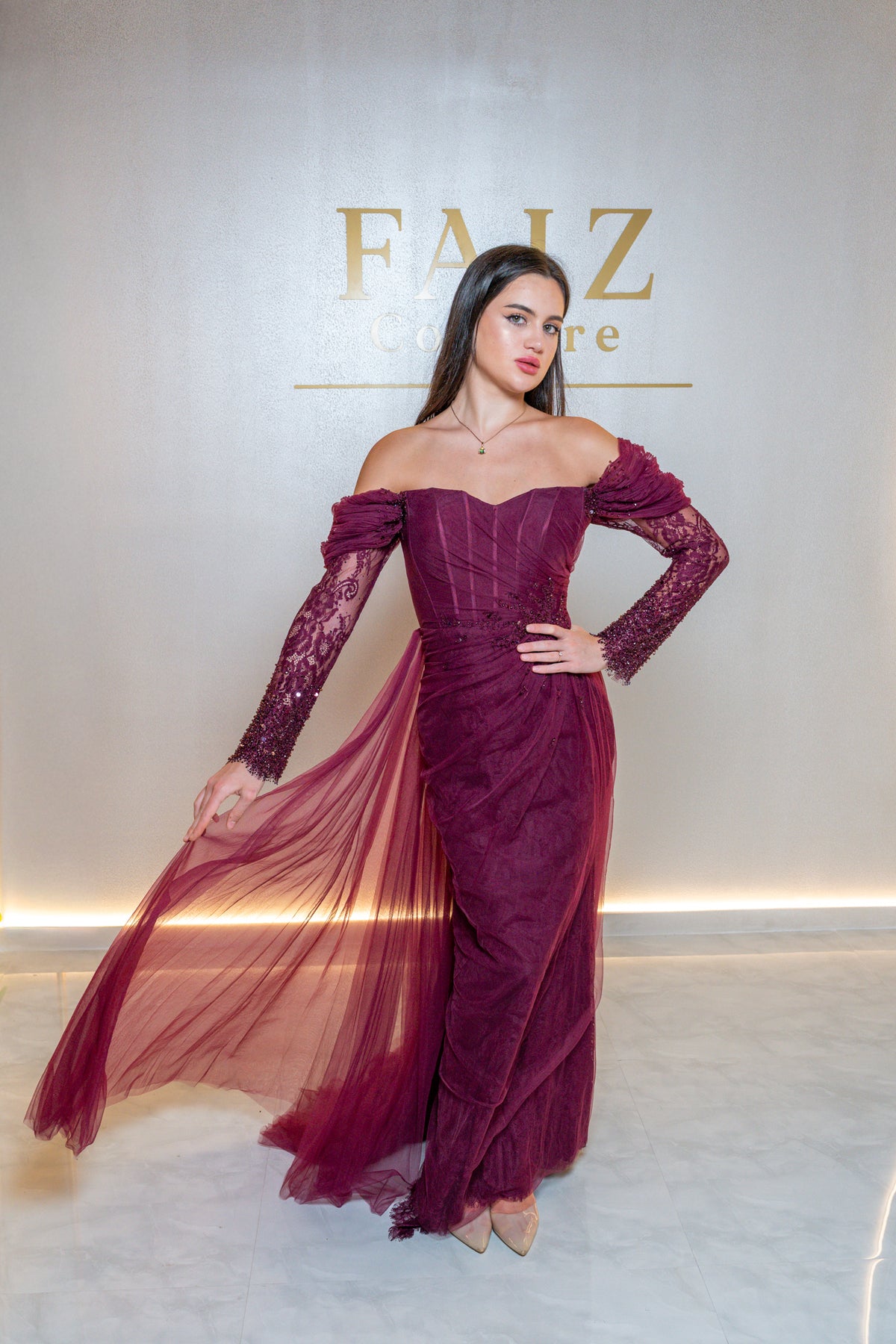 A woman models an off-the-shoulder burgundy evening gown with lace long sleeves and a flowing tulle skirt. FAIZ COUTURE