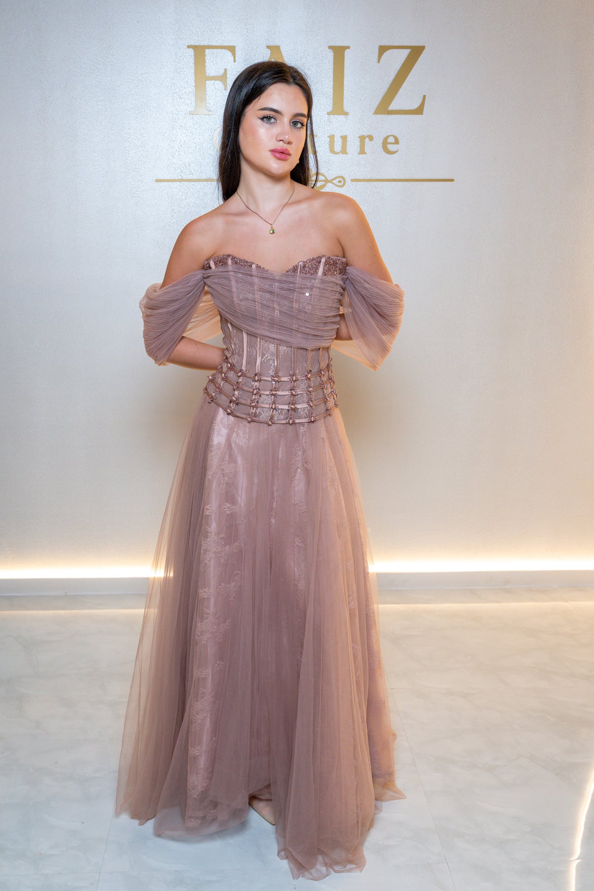 A woman modeling an off-the-shoulder floor-length gown in a dusty mauve pink color with a structured corset bodice and sheer tulle sleeves, FAIZ couture