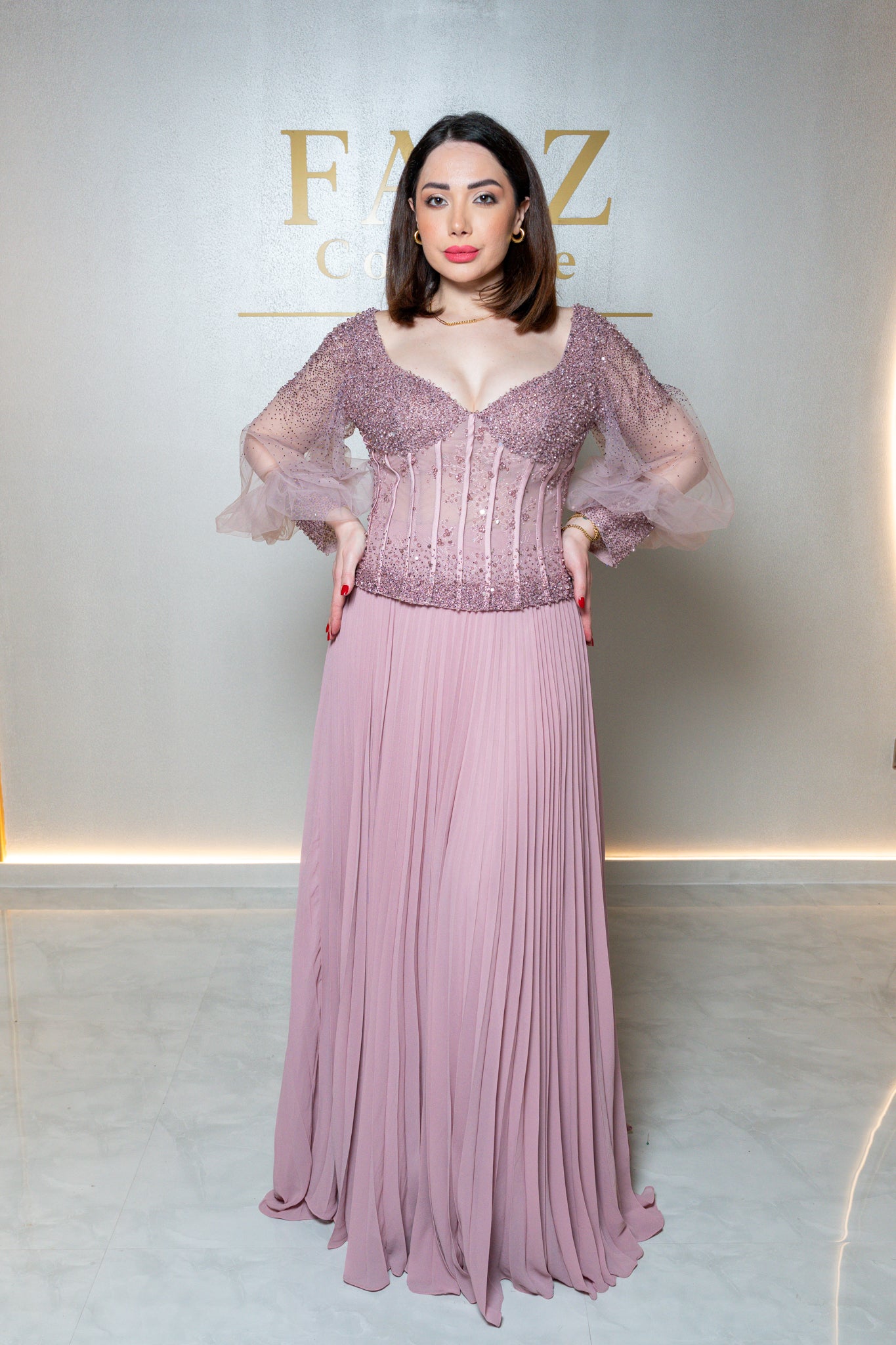 A woman models a floor-length mauve pink gown with a heavily embellished lace corset bodice and sheer puff sleeves, standing against a light gray wall with gold lettering.
