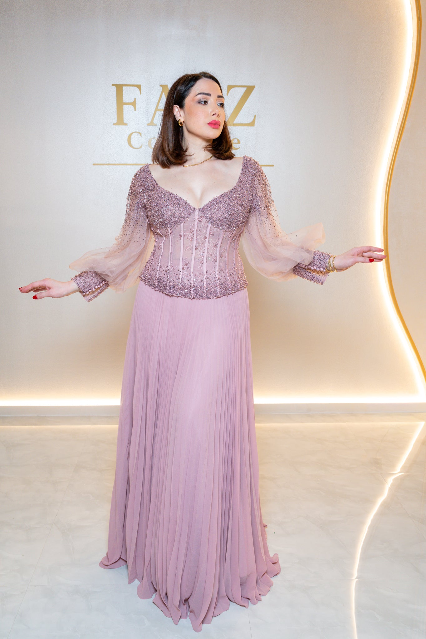 A woman modeling a floor-length lavender gown with a heavily embellished corset-style bodice and sheer flutter sleeves, posed against a warm beige wall with illuminated curves and partial gold lettering.