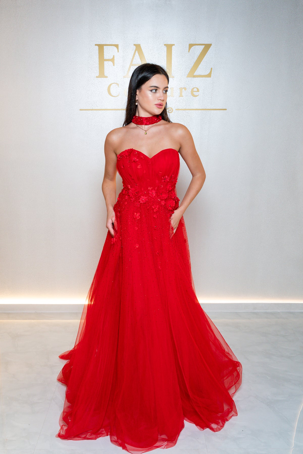 A woman modeling a bright red strapless gown with a sweetheart neckline, floral lace bodice, and flowing tulle skirt, FAIZ COUTURE