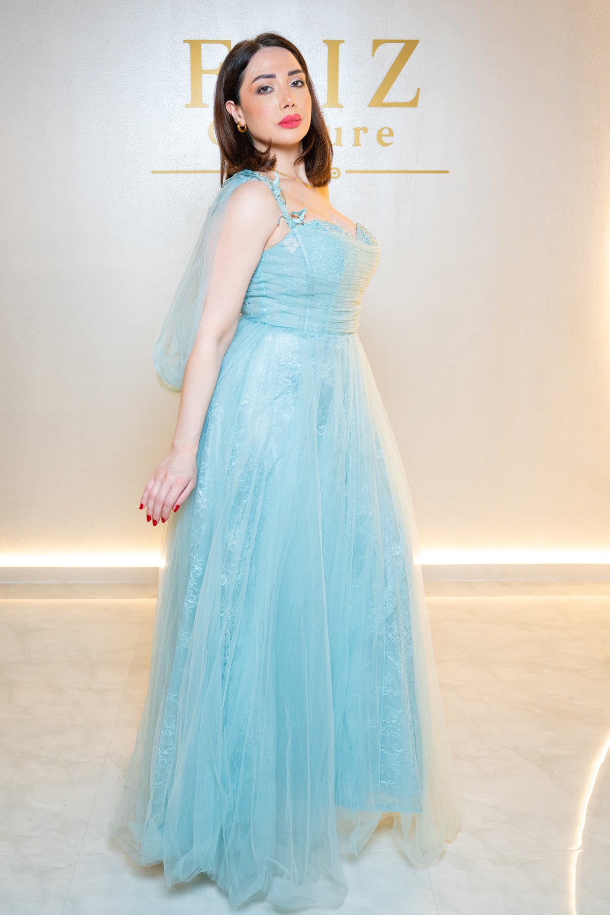 A woman wearing a light blue floor-length tulle gown with a sheer draped shoulder detail, FAIZ COUTURE