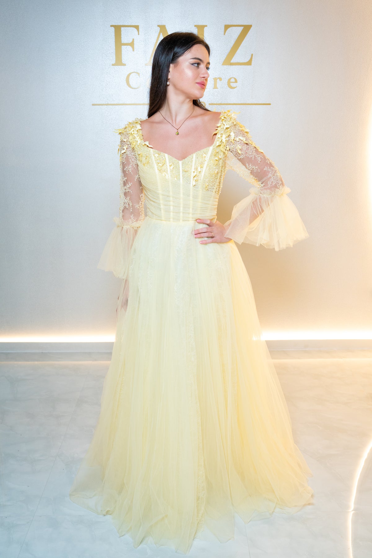 A woman modeling a pale yellow formal gown with a structured corset-style bodice, sheer lace long sleeves, and a full tulle skirt, FAIZ COUTURE