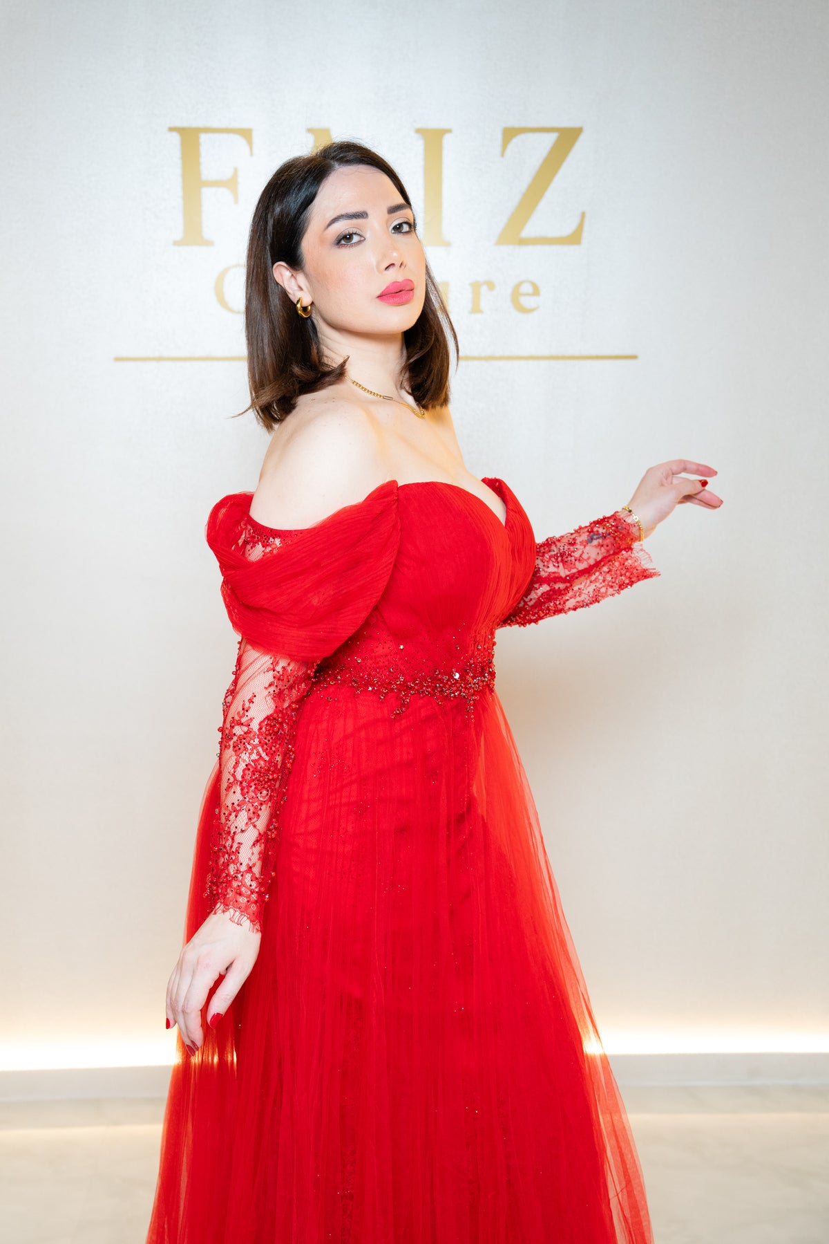 A woman wearing a vibrant red off-the-shoulder evening gown with long lace sleeves and a beaded waistband, FAIZ COUTURE