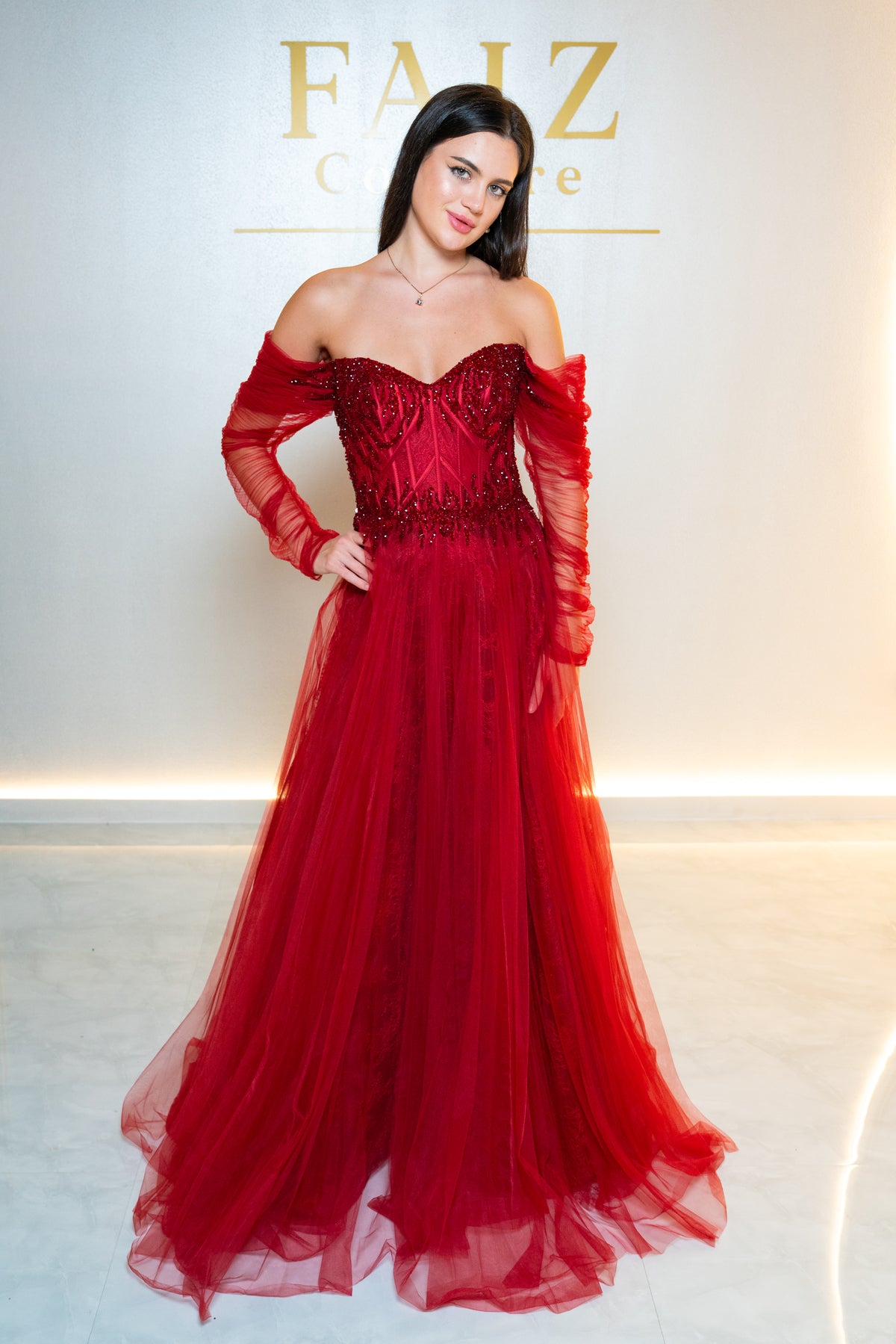 A woman modeling an off-the-shoulder red tulle gown with a structured beaded bodice and long sheer sleeves, FAIZ COUTURE