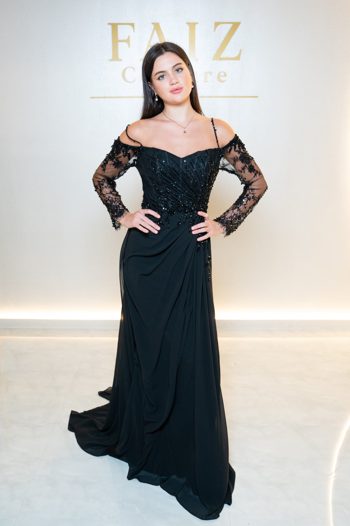 A woman modeling a black off-the-shoulder evening gown with sheer lace long sleeves and a beaded bodice, FAIZ Couture