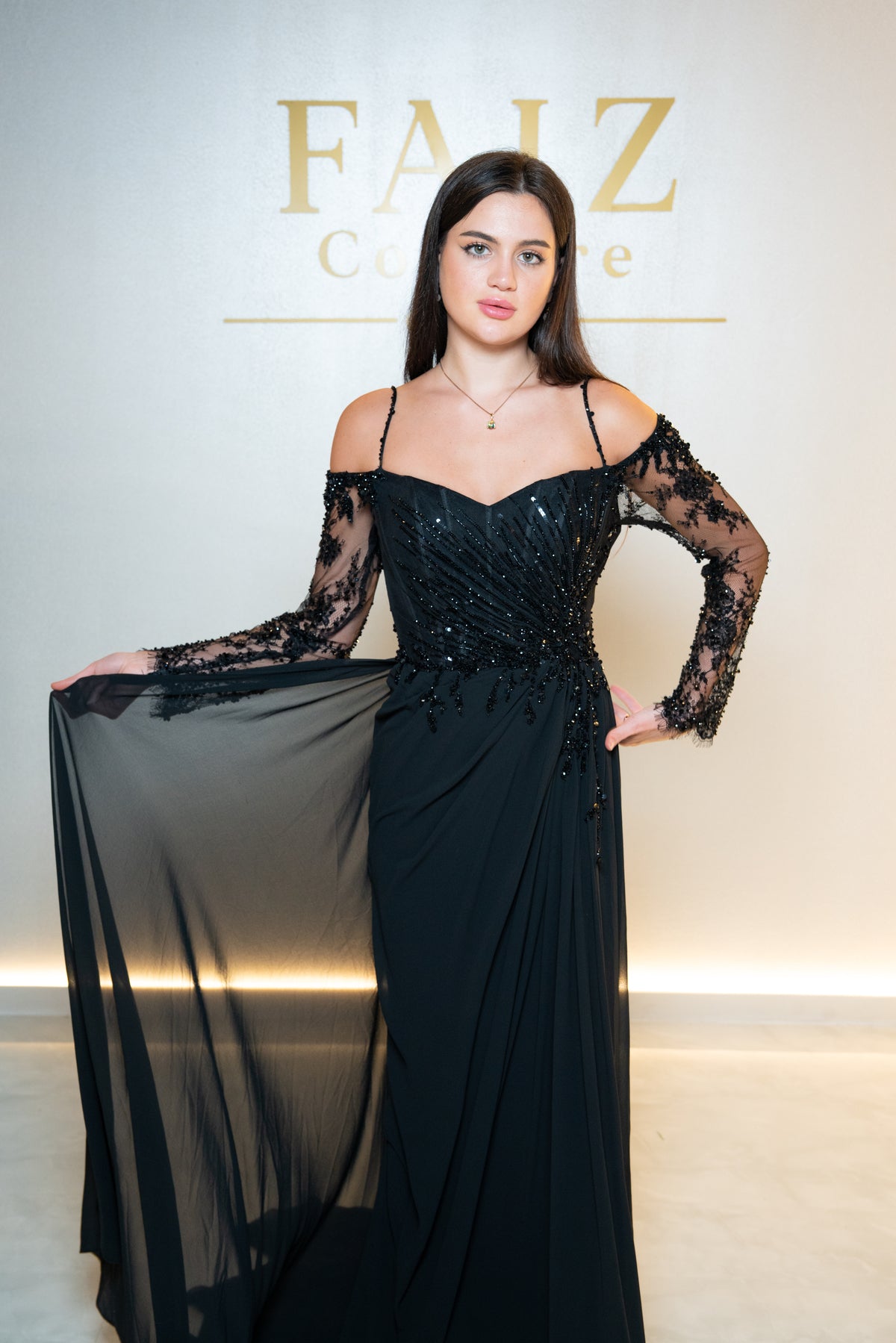 A woman modeling a black off-the-shoulder evening gown with sheer lace long sleeves and beaded embellishments, FAIZ Couture