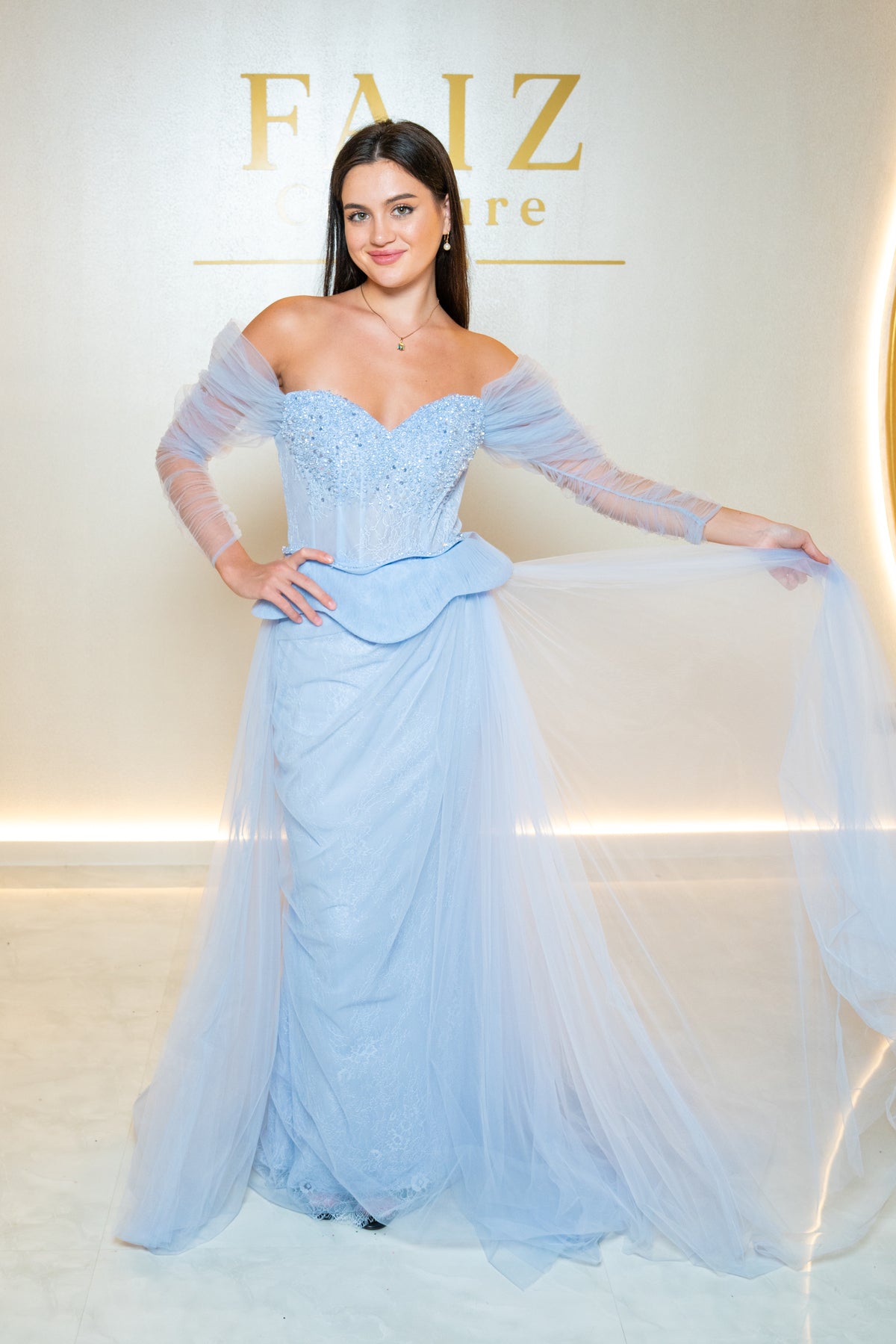A woman models a light blue off-the-shoulder gown with a structured lace bodice, sheer long sleeves, and a flowing tulle skirt, FAIZ COUTURE